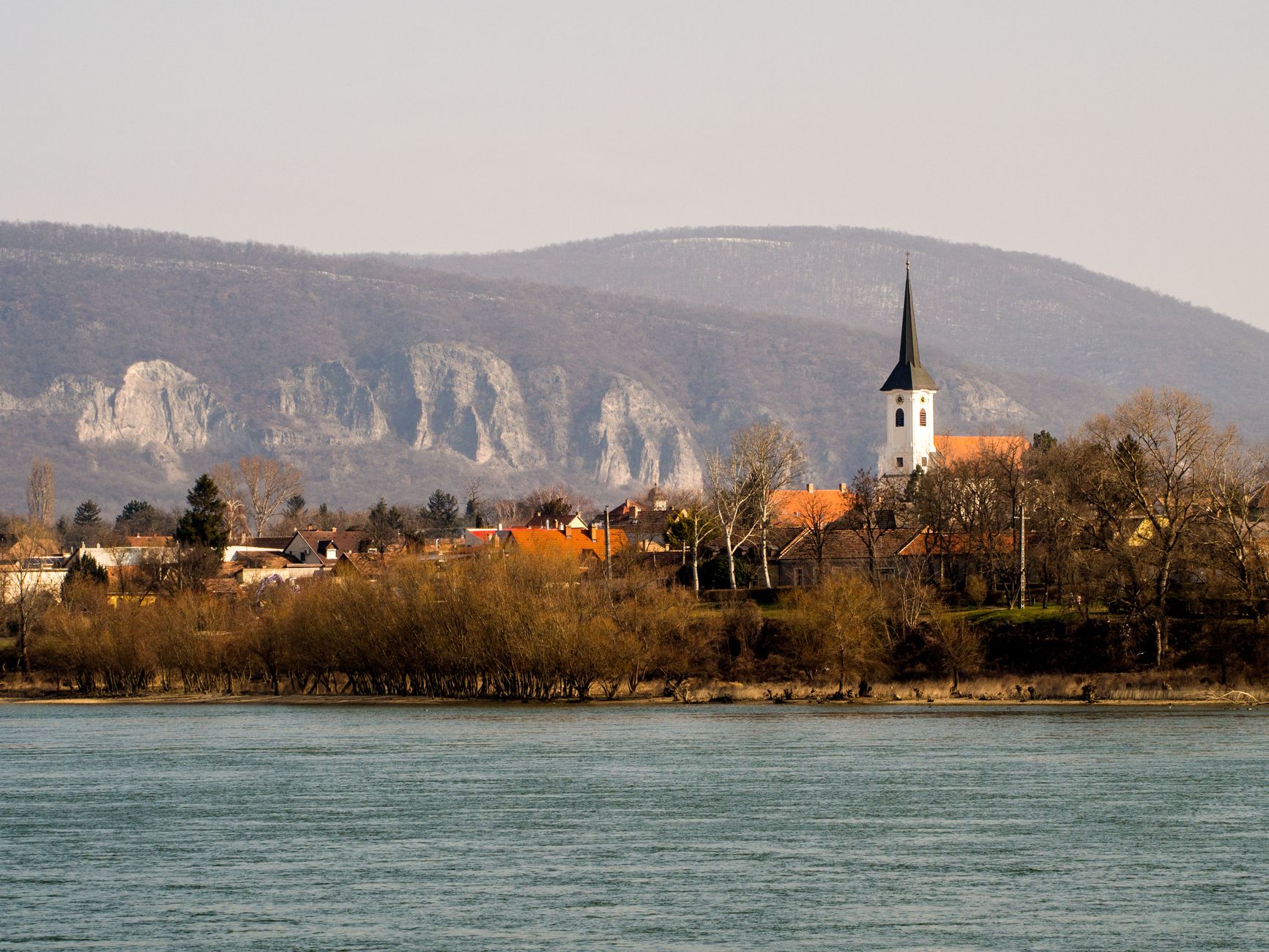 A quiet riverside village with a modest church spire nestled among green hills — evoking the un-touristed, authentic character of small Danube-side settlements that define the lower Danube experience.