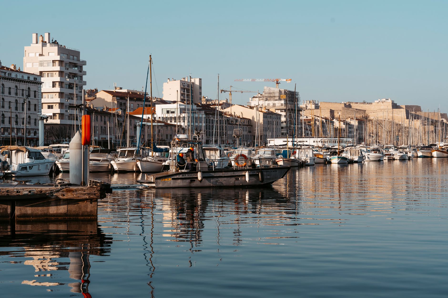 Panoramic view of Marseille's Vieux-Port harbor with sailboats moored along the quay and sunlight reflecting on the water, evoking the golden-hour ambiance of a pre-cruise overnight stay in the Mediterranean.