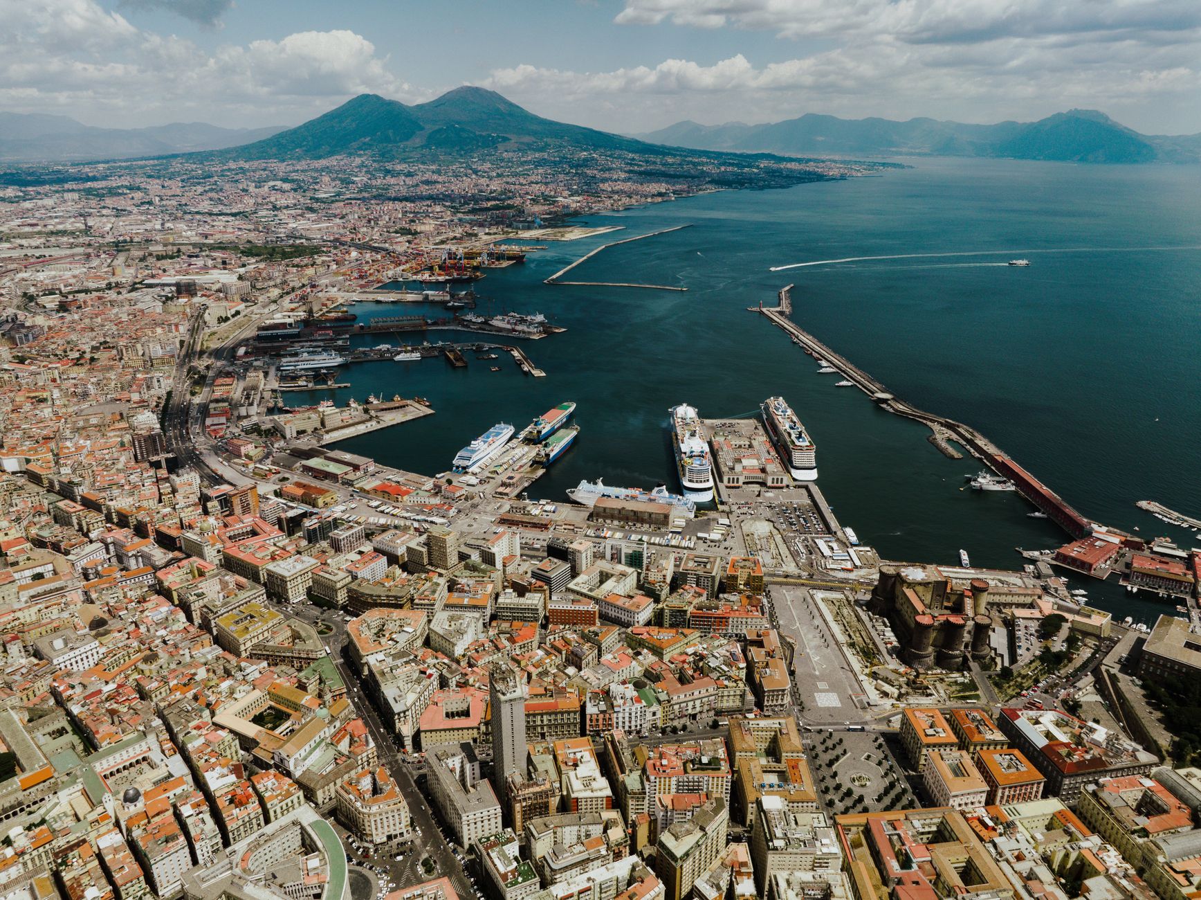 Aerial view of Naples harbor with Mount Vesuvius rising behind the city's waterfront buildings and boats moored in the bay