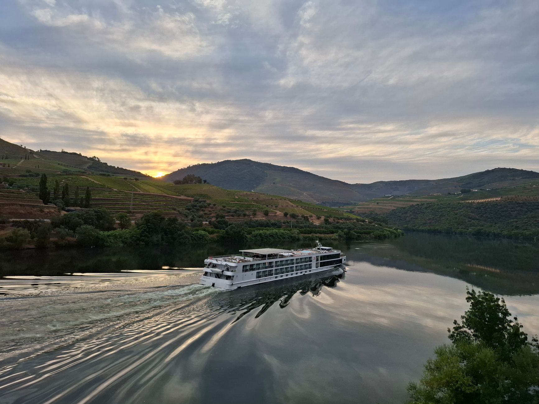 A riverboat cruising through the terraced vineyards of the Douro Valley at golden hour, capturing the immersive landscape of a Douro River cruise departing from Porto.