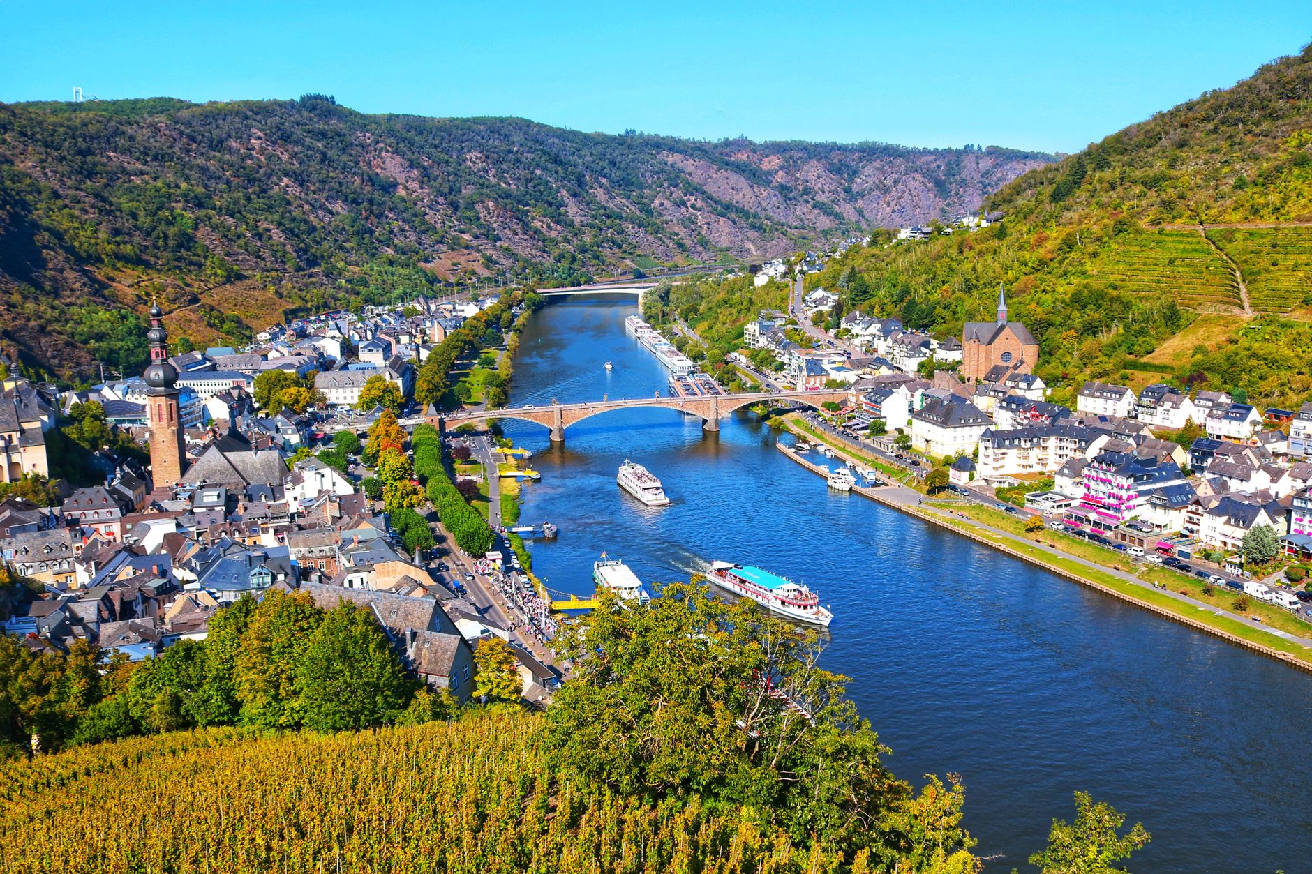 Aerial view of Cochem on the Moselle River featuring boats on the water, the riverside town, and steep hillside vineyards — a quintessential European river cruise destination.