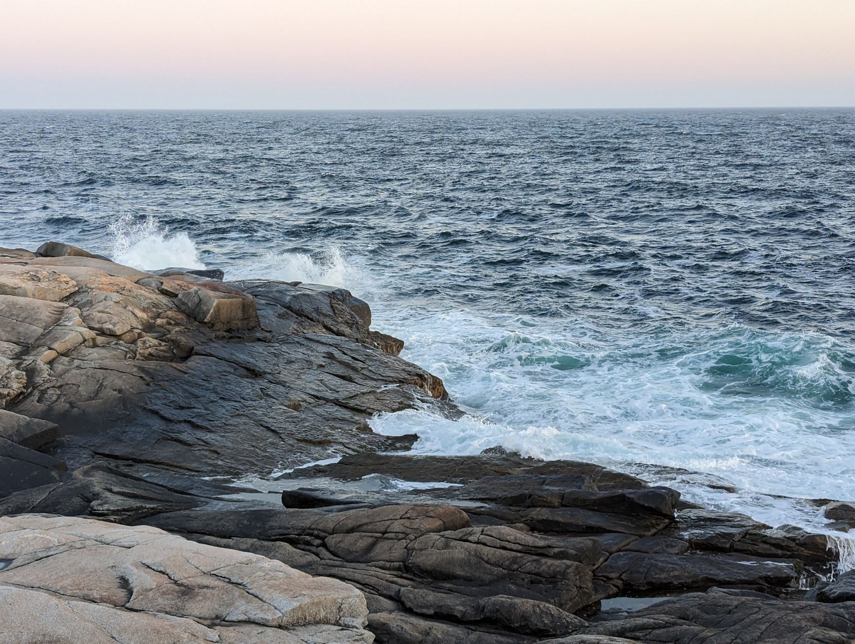 Rocky shoreline with ocean waves crashing against the granite coast at Peggy's Cove, Nova Scotia, bathed in warm sunrise light.