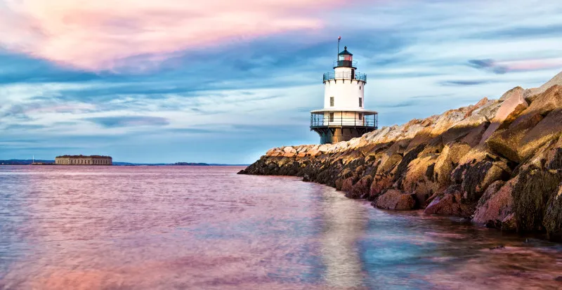 Maine lighthouse with rocky Atlantic coastline, dawn light, and crashing waves creating maritime atmosphere.
