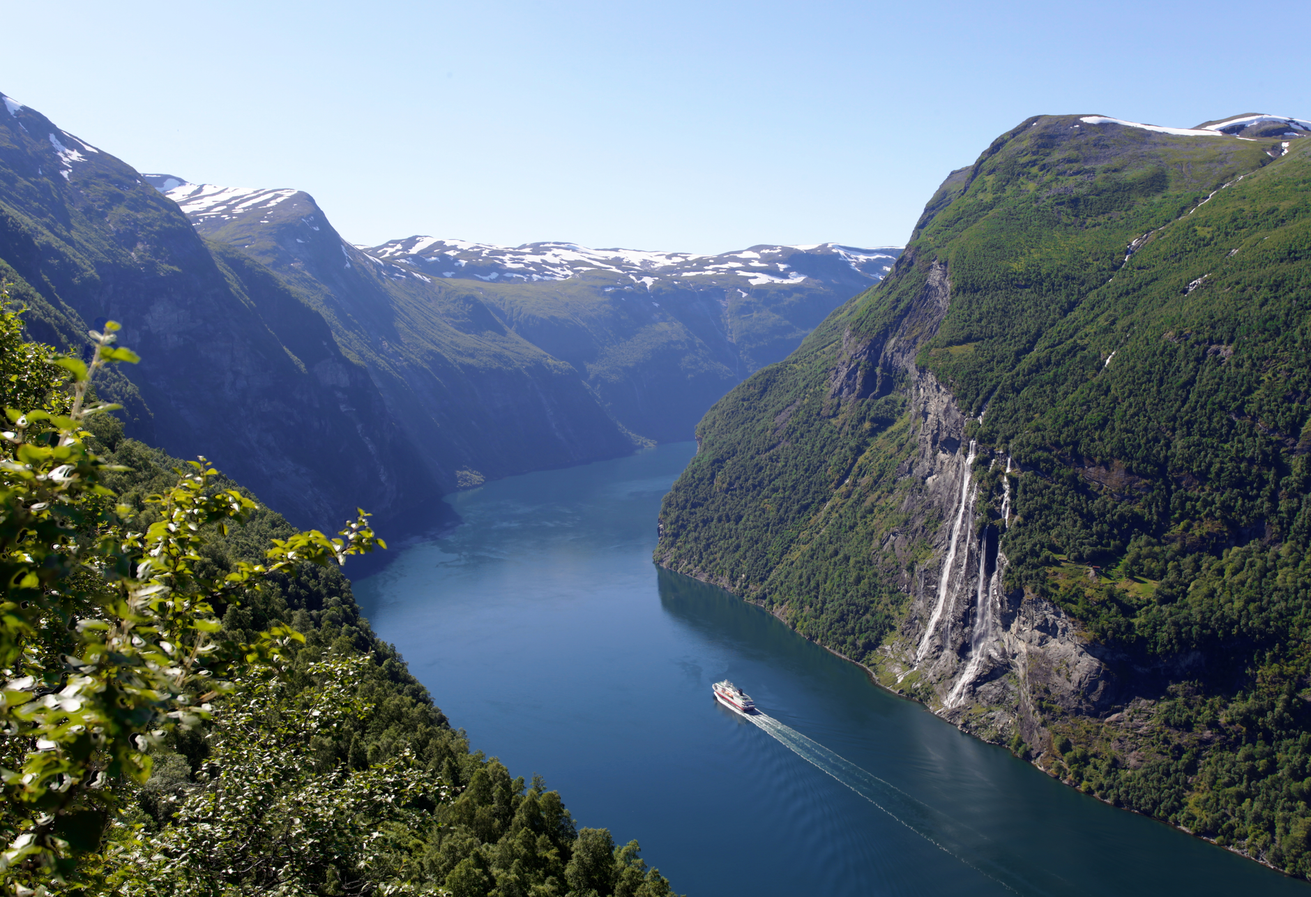 A cruise ship navigating the narrow waters of Geiranger Fjord in Norway, flanked by lush green mountainsides rising steeply from the calm water