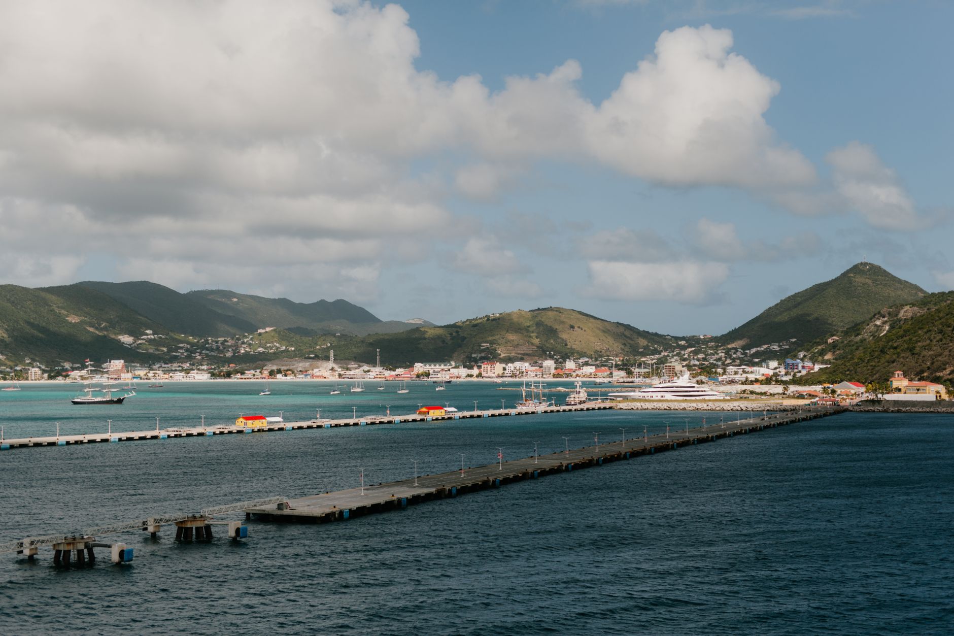 Tranquil Caribbean harbor of St. Maarten with colorful waterfront town buildings, mountain backdrop, and bright tropical skies — a popular transatlantic cruise destination from Barcelona.