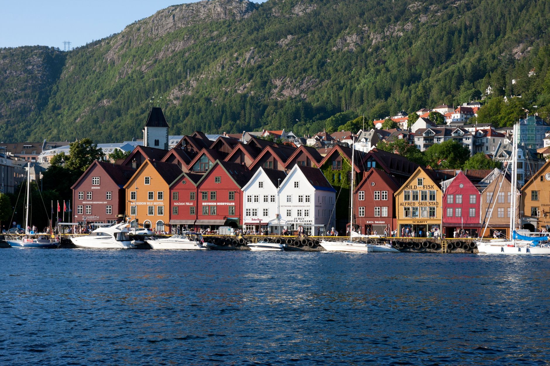 Bergen's iconic Bryggen waterfront with its colourful historic wooden buildings lining the harbour, framed by lush green mountains — the classic departure view for cruise passengers leaving the city.