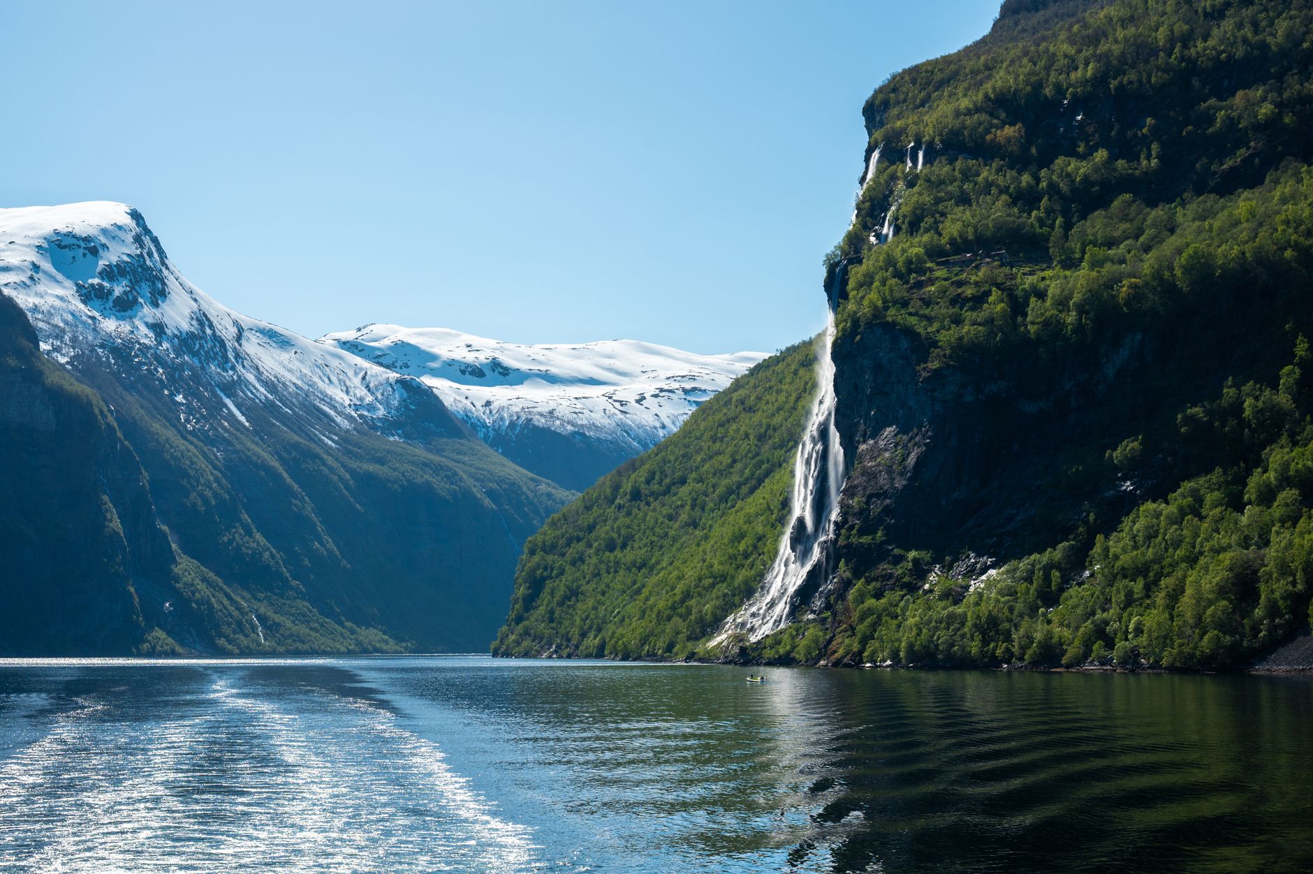 Geirangerfjord in Norway with a cascading waterfall plunging down steep rock faces into the fjord, framed by snow-capped mountain peaks and atmospheric clouds