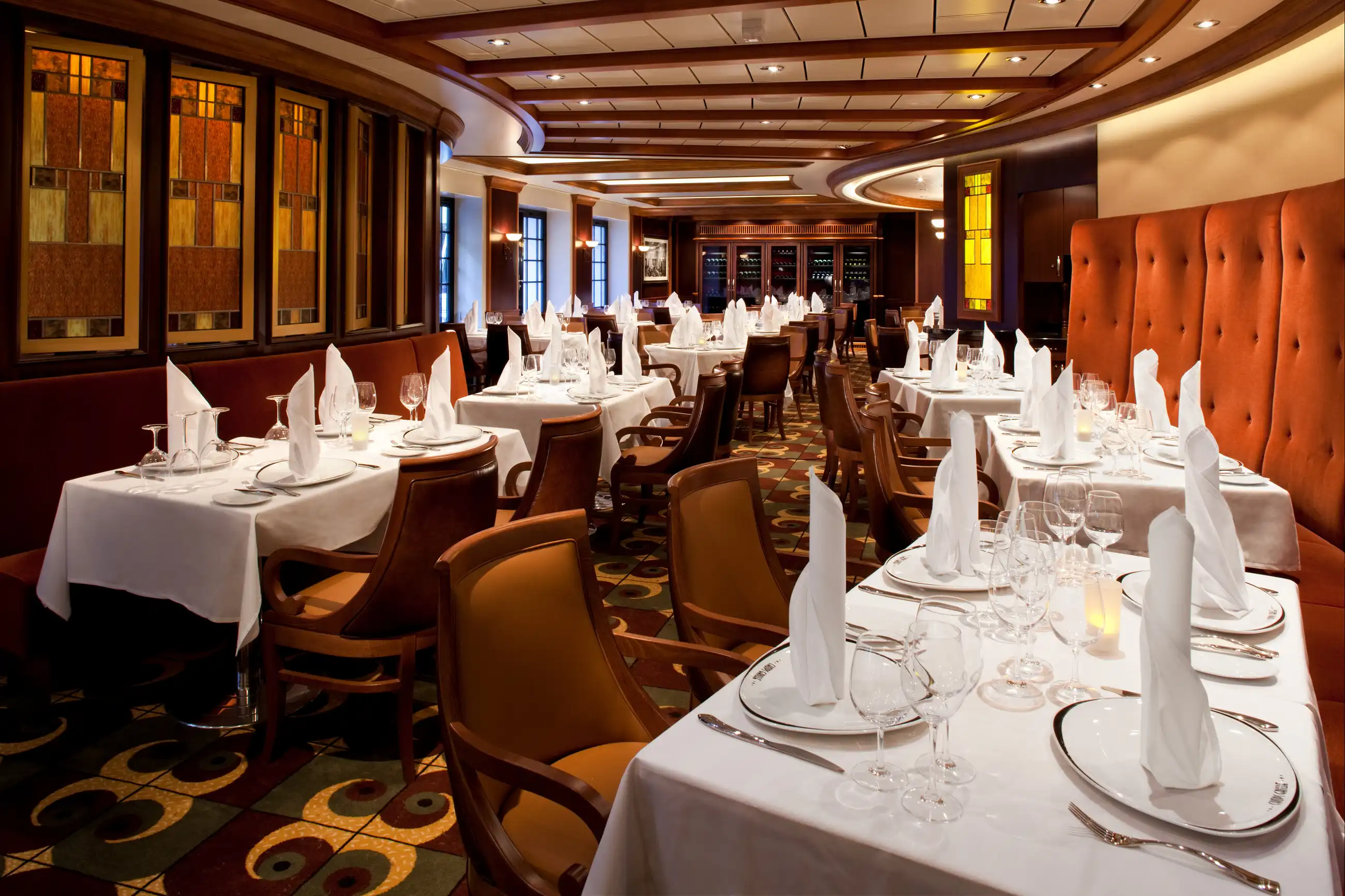 The Chops Grille steakhouse dining room aboard Allure of the Seas, with wood-paneled walls, burnt-orange tufted banquettes and white-draped tables set for dinner.
