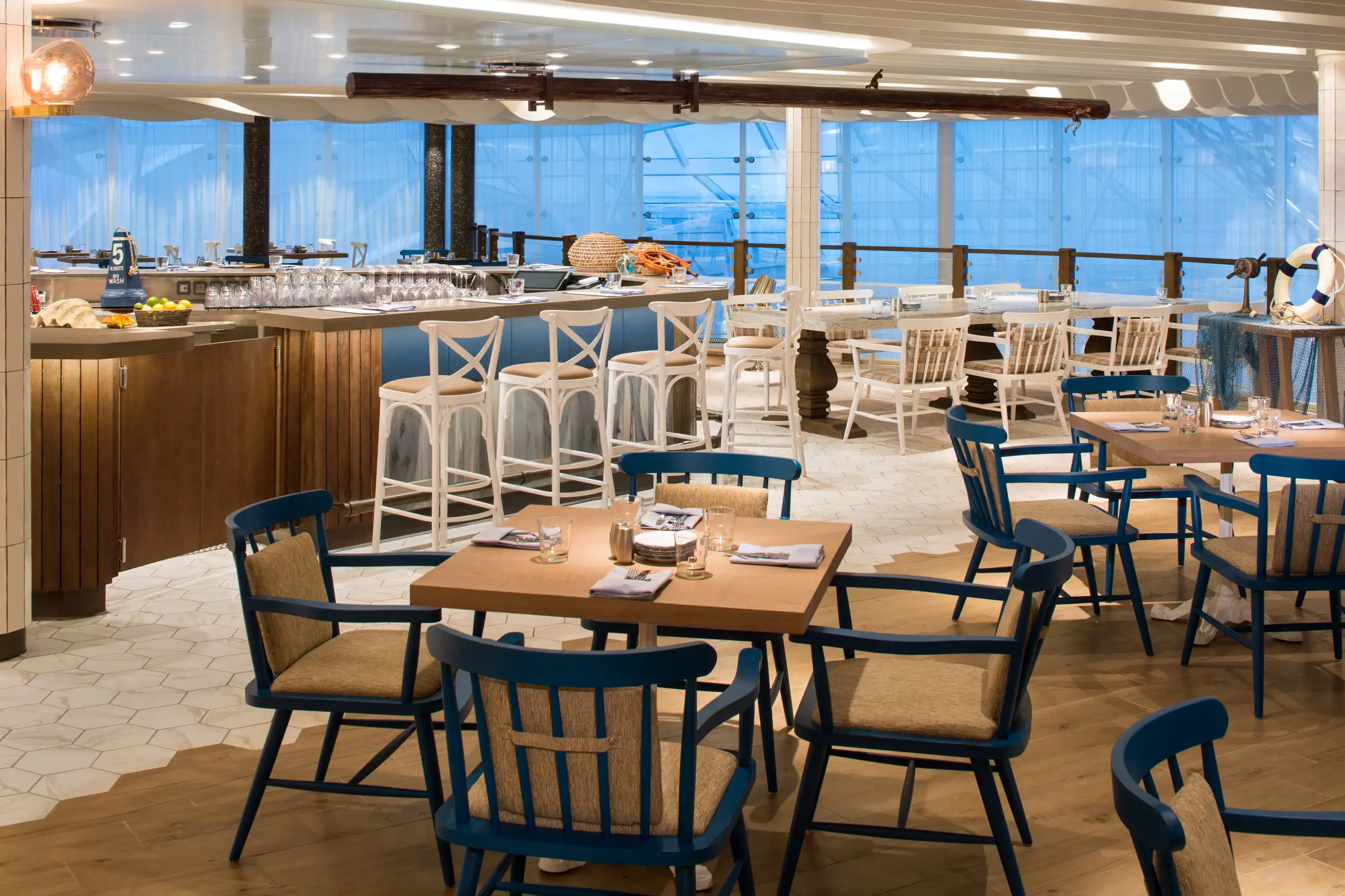 The empty Hooked Seafood dining room on Symphony of the Seas, with wooden tables, navy chairs, a white-stooled bar and floor-to-ceiling ocean windows.