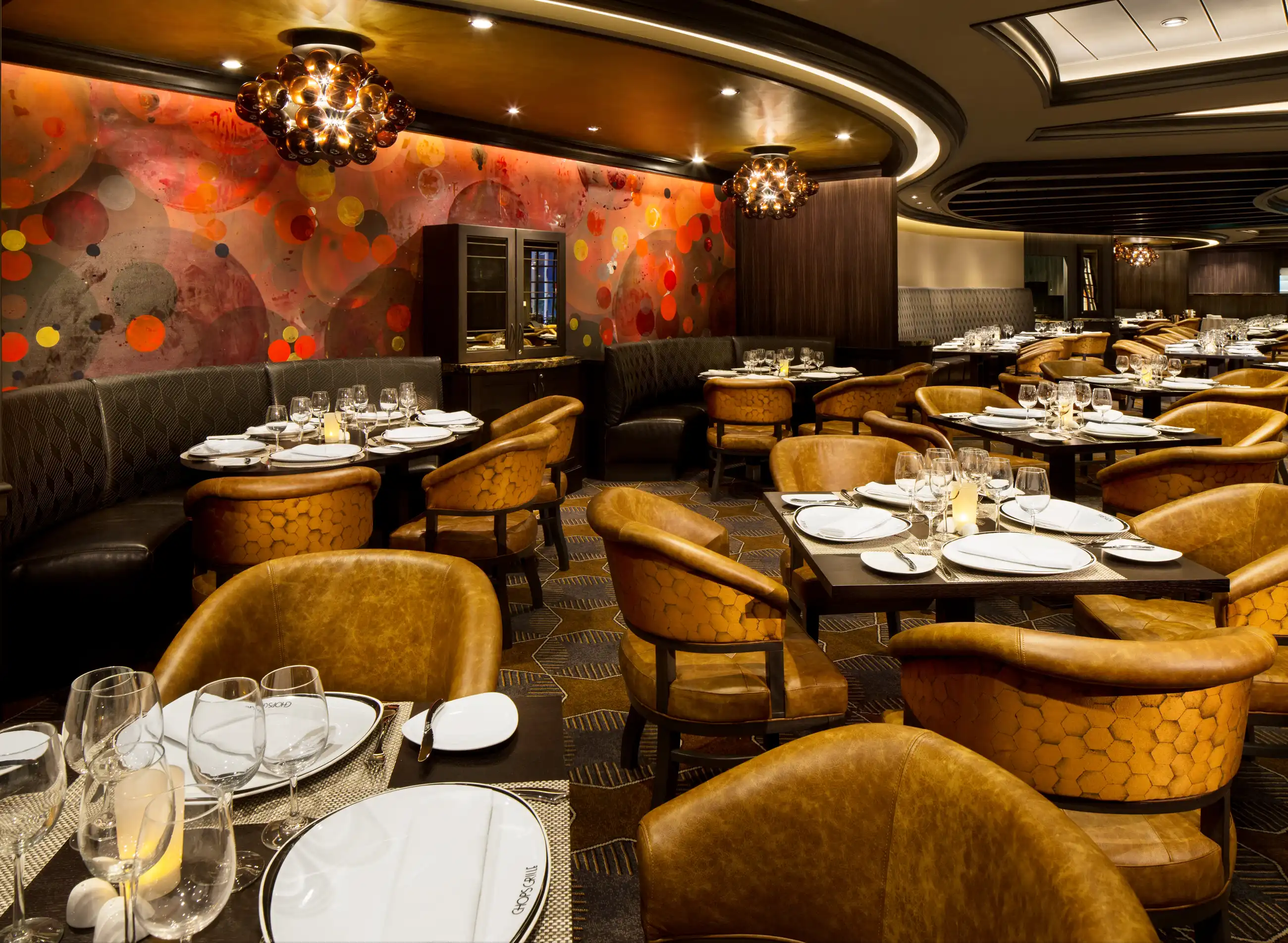 Chops Grille aboard Harmony of the Seas, with mustard-gold curved banquettes, warm wood paneling and white-clothed tables beneath abstract red and orange artwork.