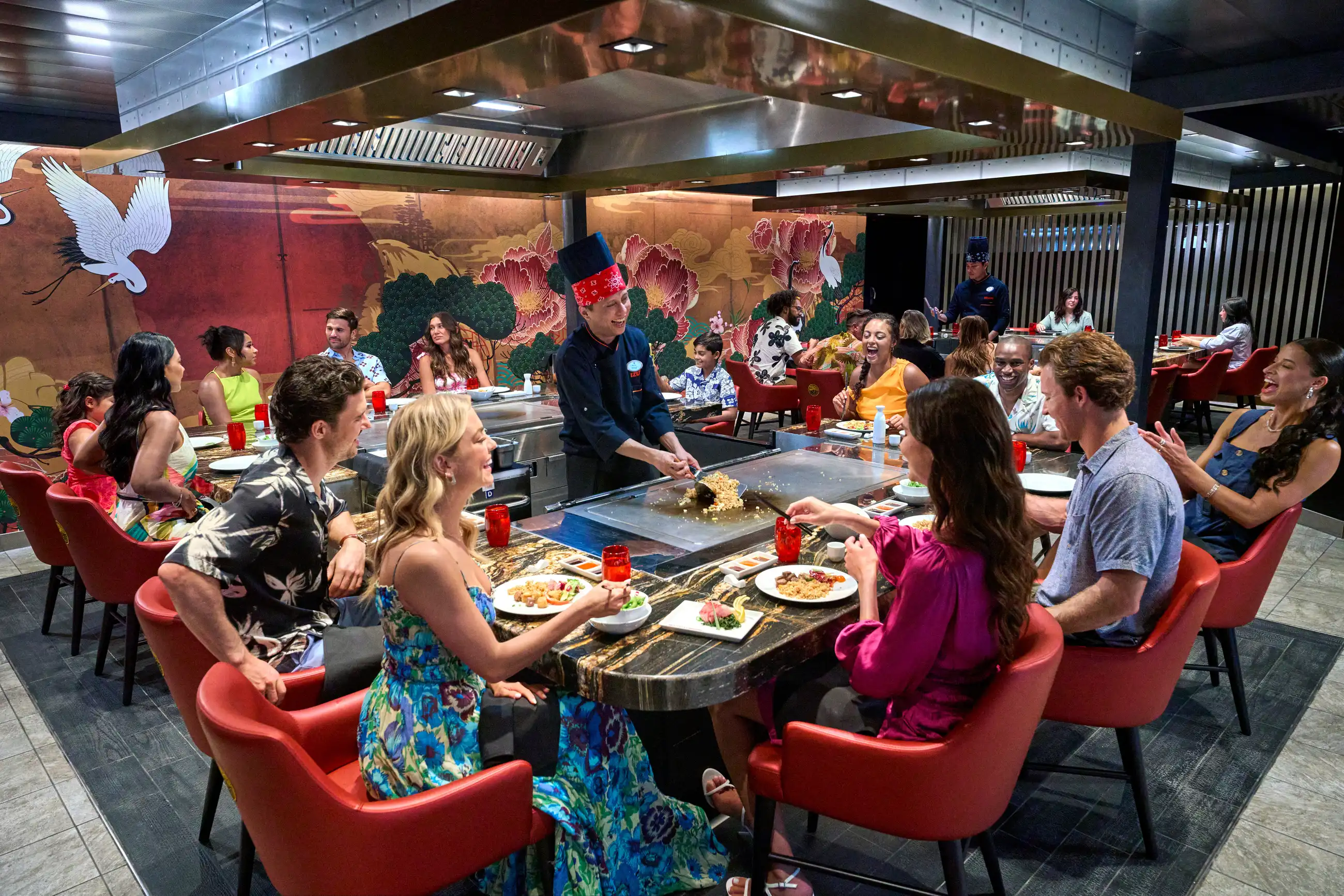 Guests seated around teppanyaki griddle tables at Izumi Hibachi on Utopia of the Seas while a chef cooks, with red chairs and floral wall murals.
