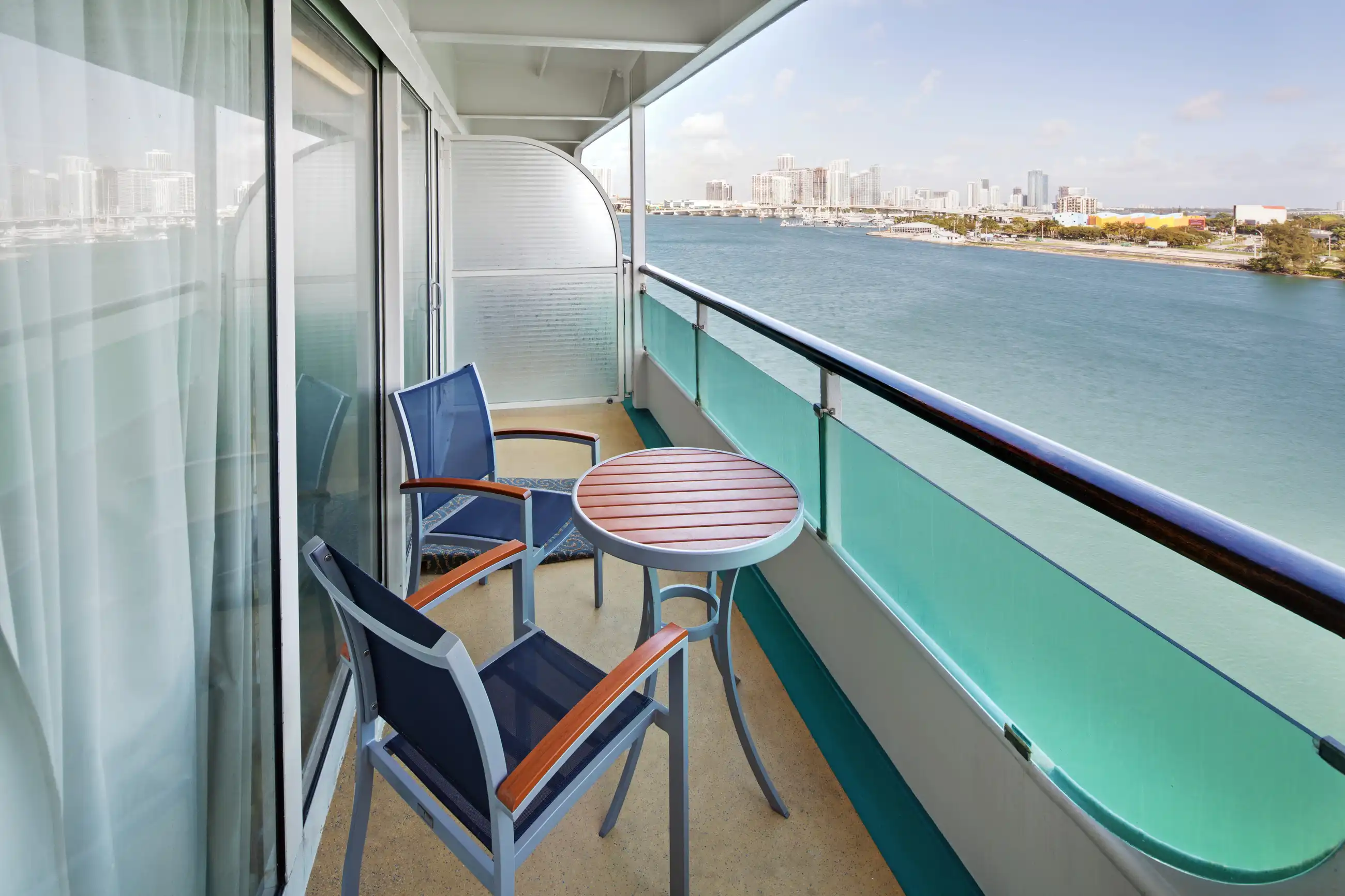 A private ocean-view balcony cabin with a small round table and two chairs, overlooking turquoise waters with a distant coastal city skyline and beachfront visible across the waterway. The cabin features floor-to-ceiling glass doors, modern furnishings, and the typical rail design of a cruise ship stateroom.