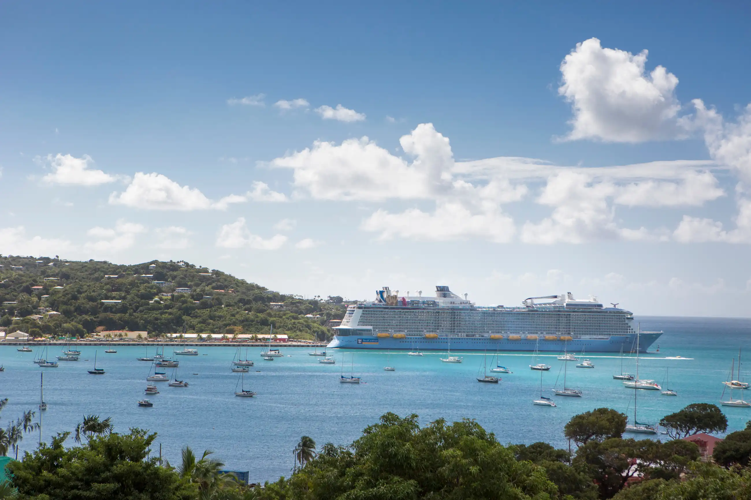 Royal Caribbean's Quantum of the Seas, one of the two ships in the Singapore programme, at anchor in turquoise water off Charlotte Amalie harbour, St. Thomas, with sailboats and green hills behind her.
