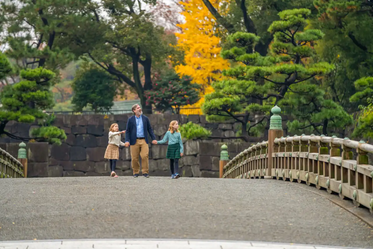 An adult and two children cross a traditional arched stone bridge in a manicured Japanese garden with evergreens and autumn foliage.