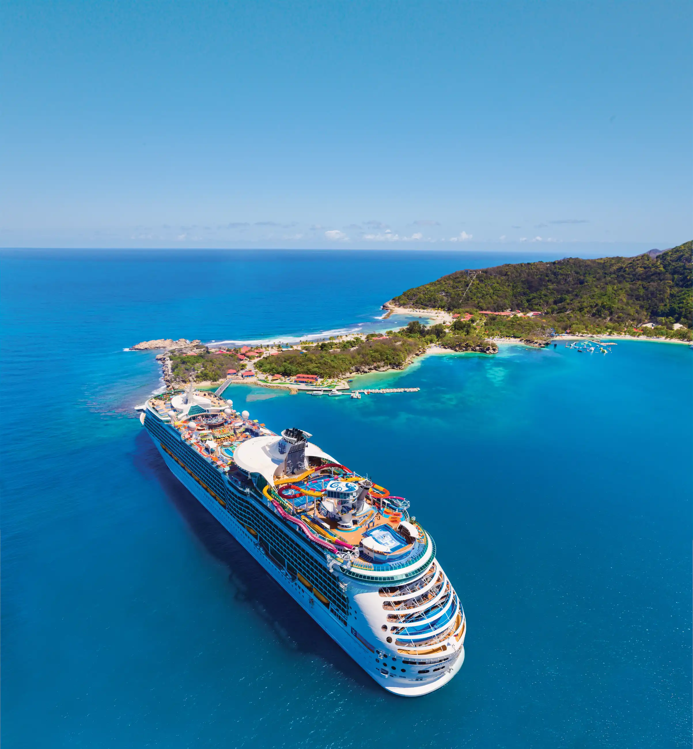 Aerial view of Royal Caribbean's Navigator of the Seas anchored in turquoise Caribbean water beside a forested, beach-lined coastline.