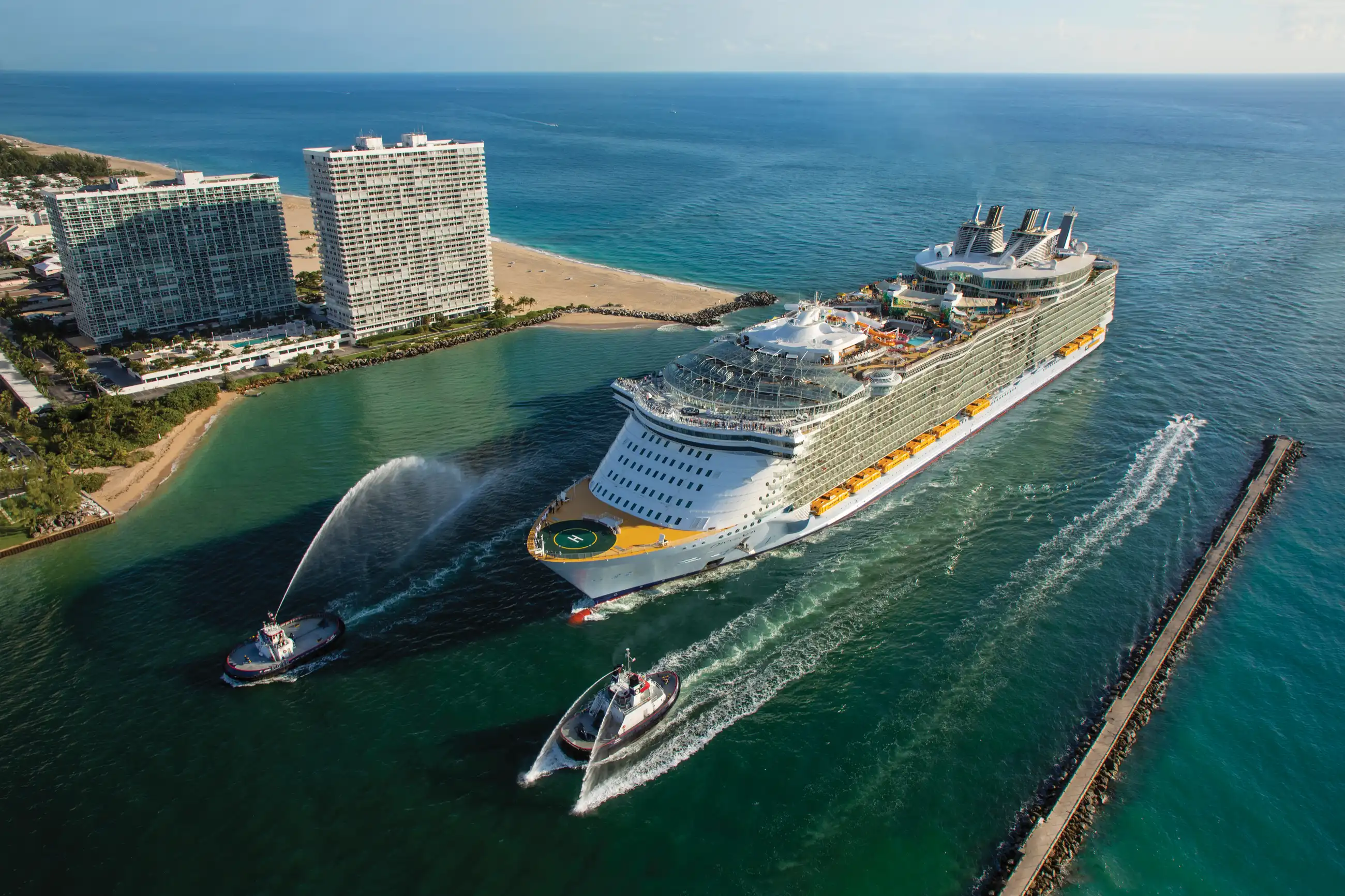 Aerial view of a Royal Caribbean Oasis-class ship berthed at Port Everglades, Fort Lauderdale, with tugboats alongside and beachfront hotels behind.
