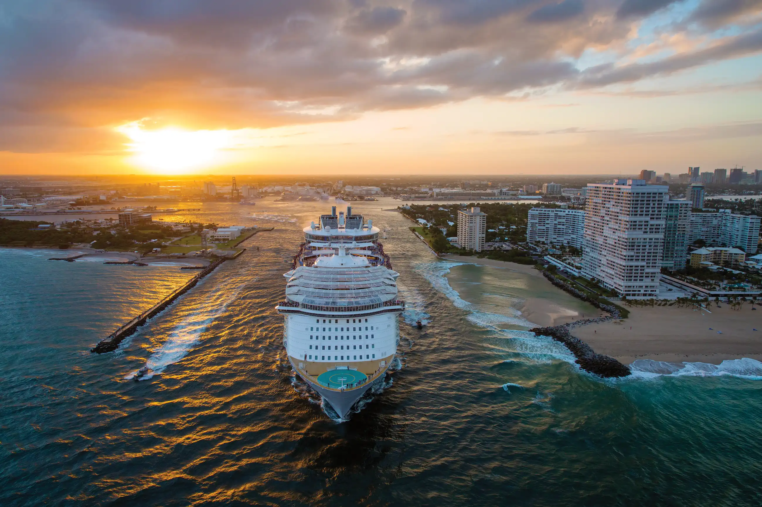 Aerial view of Royal Caribbean's Harmony of the Seas heading out through the Fort Lauderdale shipping channel at sunset, with the port city and beach on the right.
