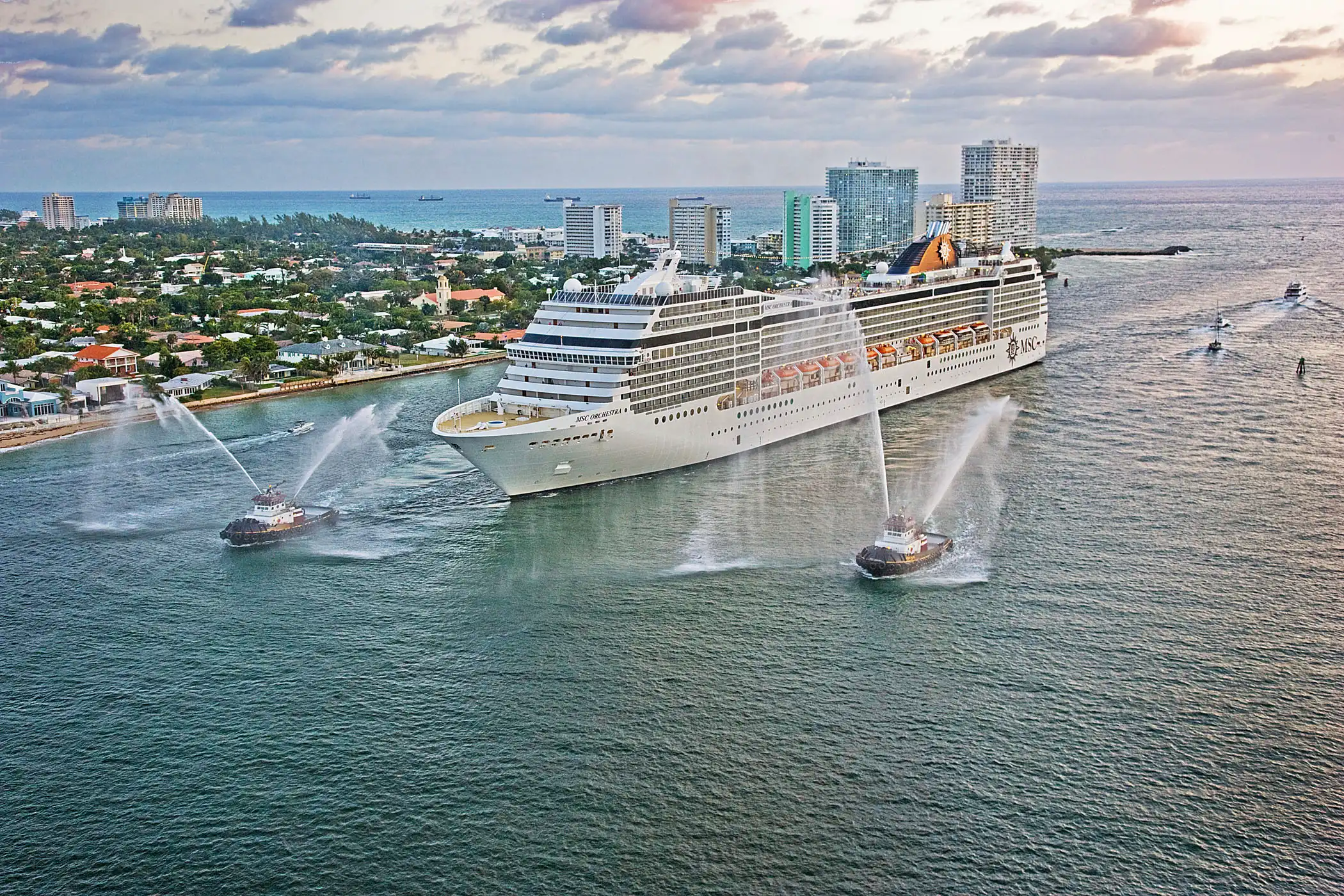 Aerial view of MSC Orchestra leaving Port Everglades at Fort Lauderdale, escorted by tugboats spraying ceremonial water arcs, with the coastal skyline behind.