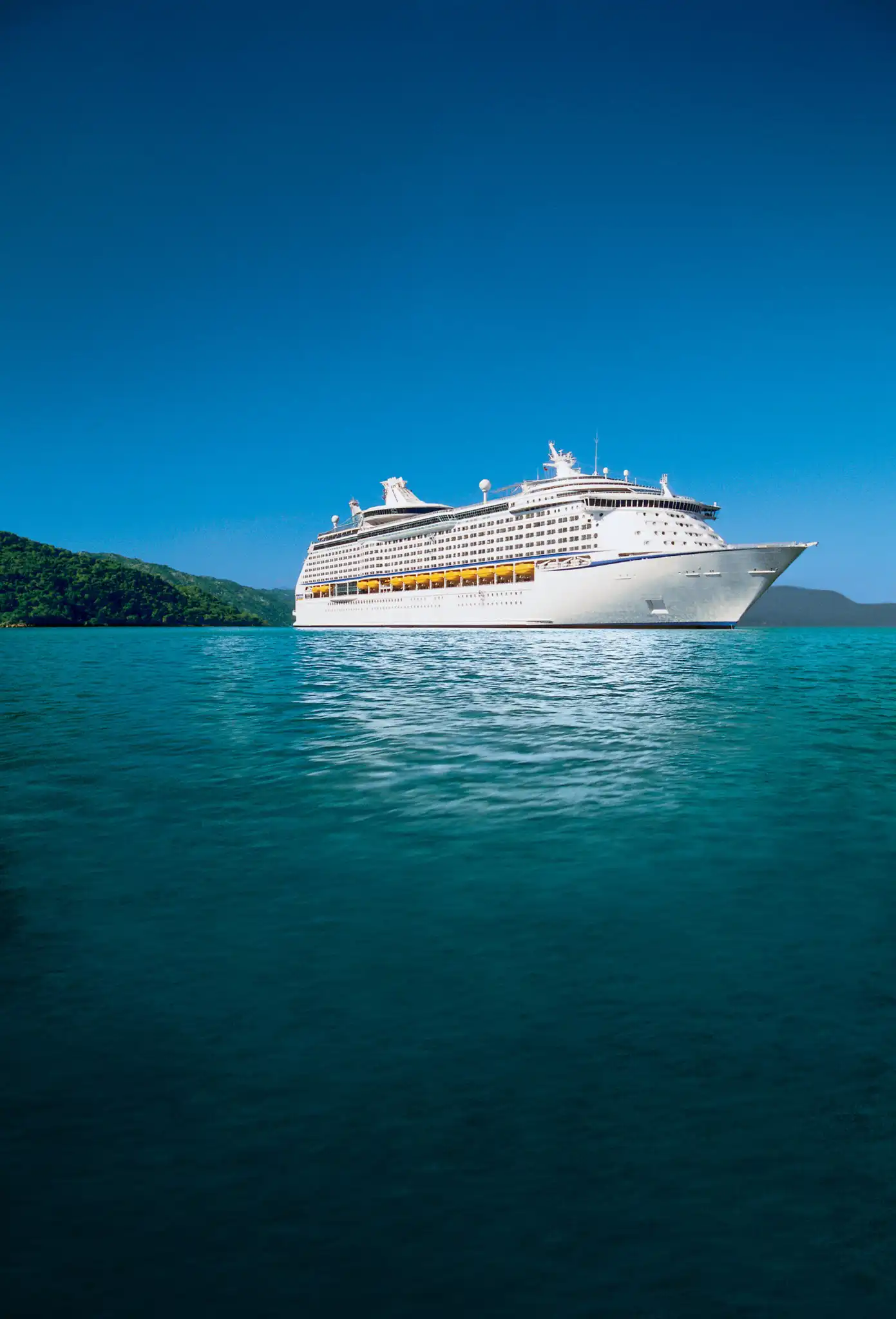 Explorer of the Seas at anchor in clear turquoise water off a forested coastline, seen from water level.