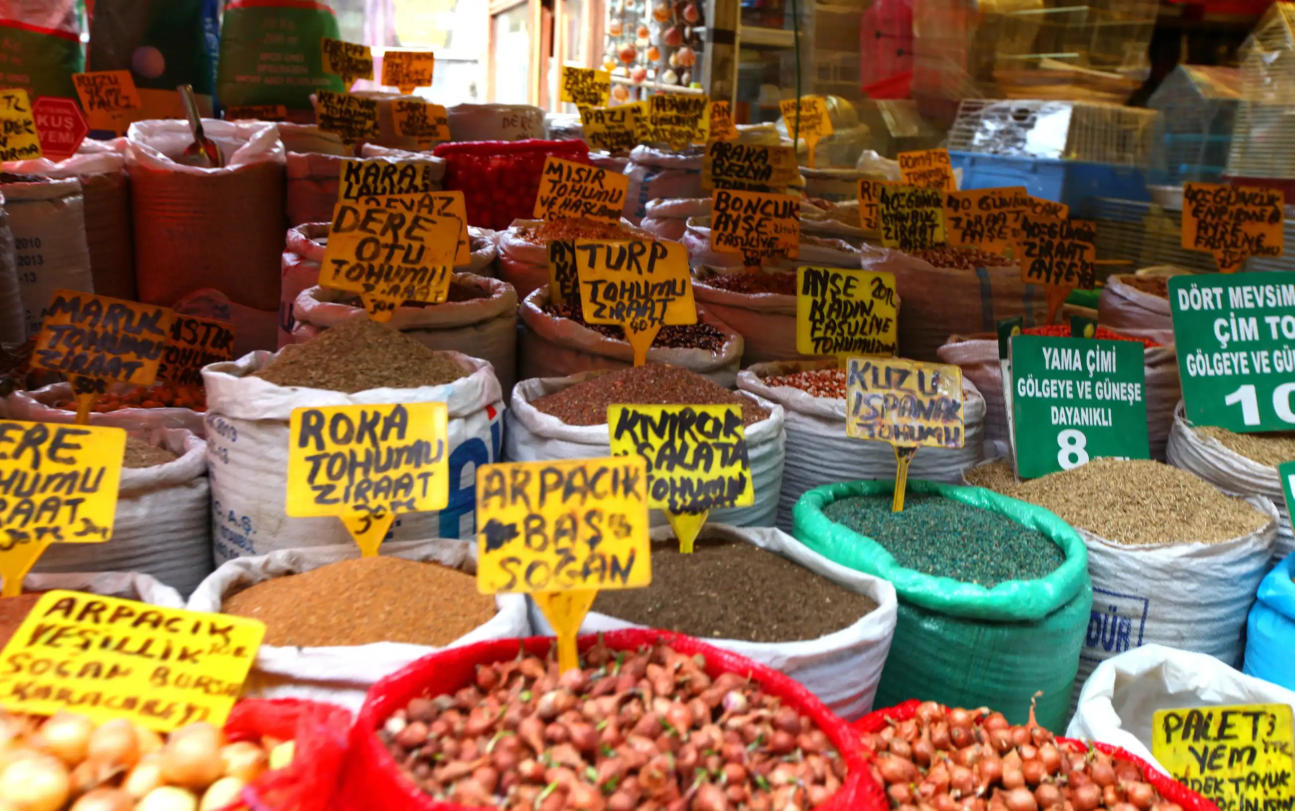 A spice market stall in a Turkish bazaar with dried spices, herbs and legumes in labelled containers and red bowls of grains.