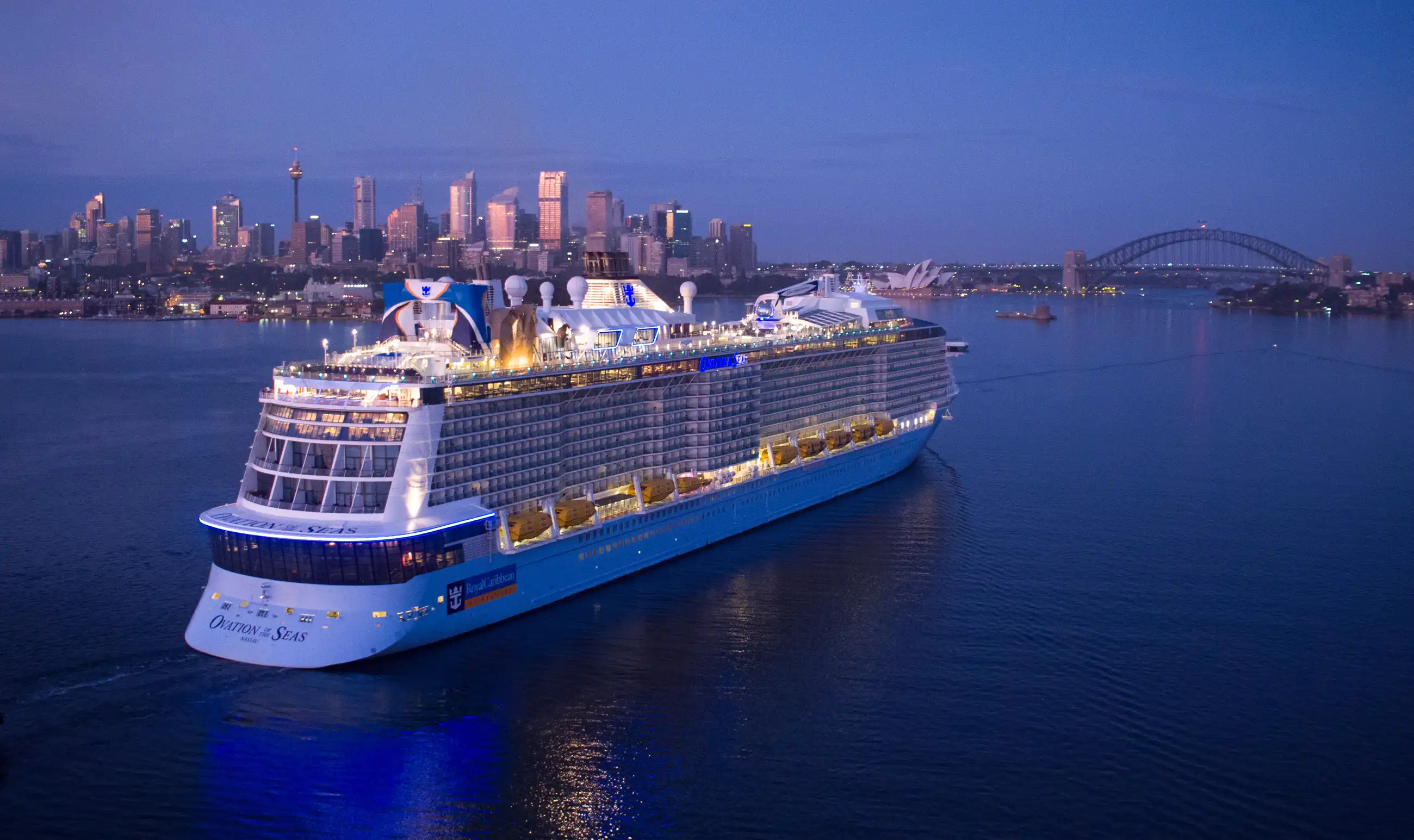 Royal Caribbean's Ovation of the Seas lit up at dusk in Sydney Harbour, with the city skyline and harbour bridge across the water.