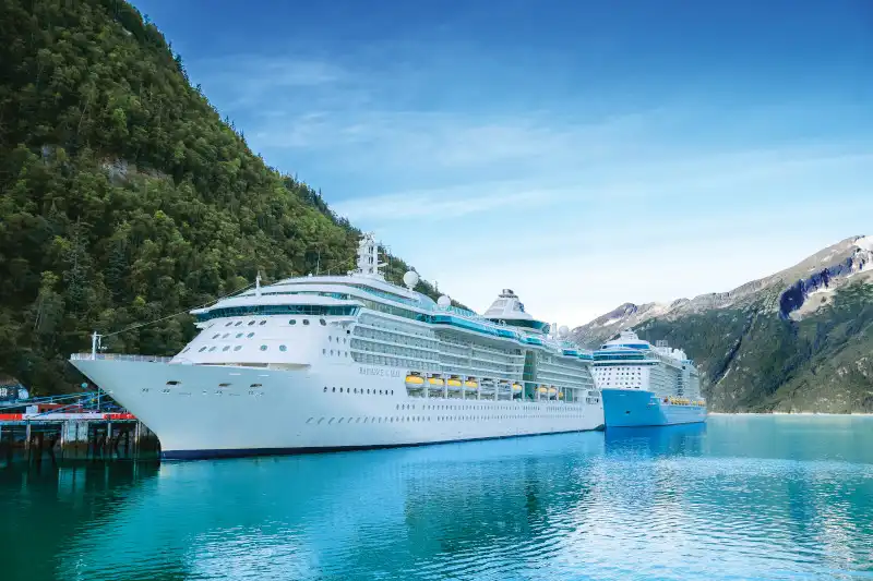 Large Royal Caribbean cruise ship docked at Skagway port in a pristine turquoise fjord surrounded by dense evergreen forest and steep mountain terrain. A second RCI ship is visible moored nearby, showcasing the scale of cruise traffic in this Alaskan destination.