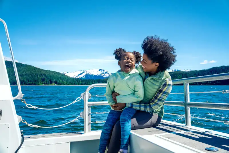 An adult and child sit together on a cruise ship's exterior deck railing, enjoying a scenic mountain and lake view. Snow-capped peaks, forested shorelines, and brilliant blue waters frame this candid moment of joy and bonding.