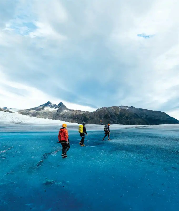 Three adventure tourists in colorful cold-weather gear hike across a brilliant blue glacial surface with a snow-covered mountain peak and rocky ridges in the background under partly cloudy skies. This is an active glacier excursion experience showing the dramatic landscape of Southeast Alaska.