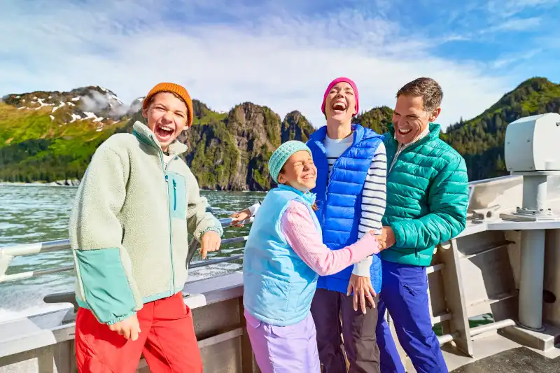 A family of four laughs together on the deck of a wildlife tour boat in Kenai Fjords, Alaska, with dramatic rocky peaks and forested shoreline visible behind them under partly cloudy skies.