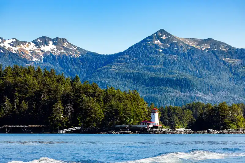 Coastal Alaskan landscape featuring snow-capped mountains rising behind a dense evergreen forest, with a white lighthouse structure visible on the forested shoreline overlooking calm blue water. This scenic cruise destination represents typical Inside Passage scenery with dramatic mountain peaks and pristine waterfront.
