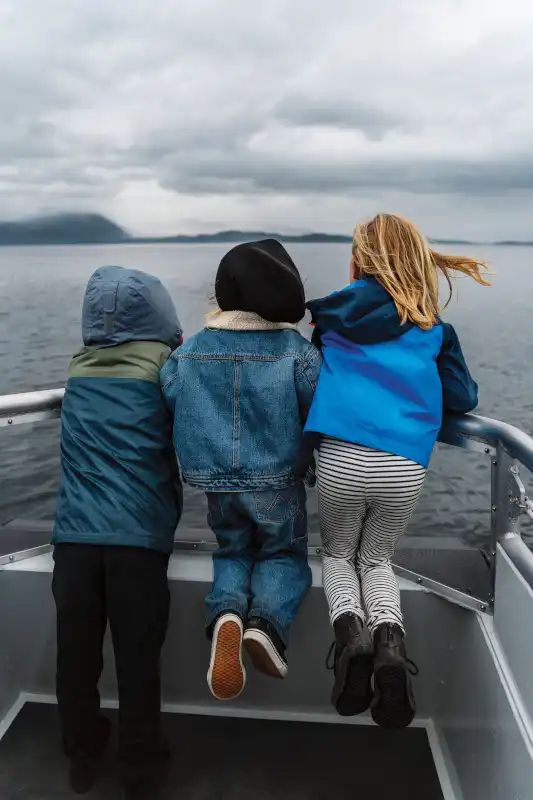 Three children seated at the railing of a boat or small vessel, viewed from behind as they gaze toward a mountainous coastal landscape. The scene captures a family excursion on calm waters under overcast skies with distant islands visible.