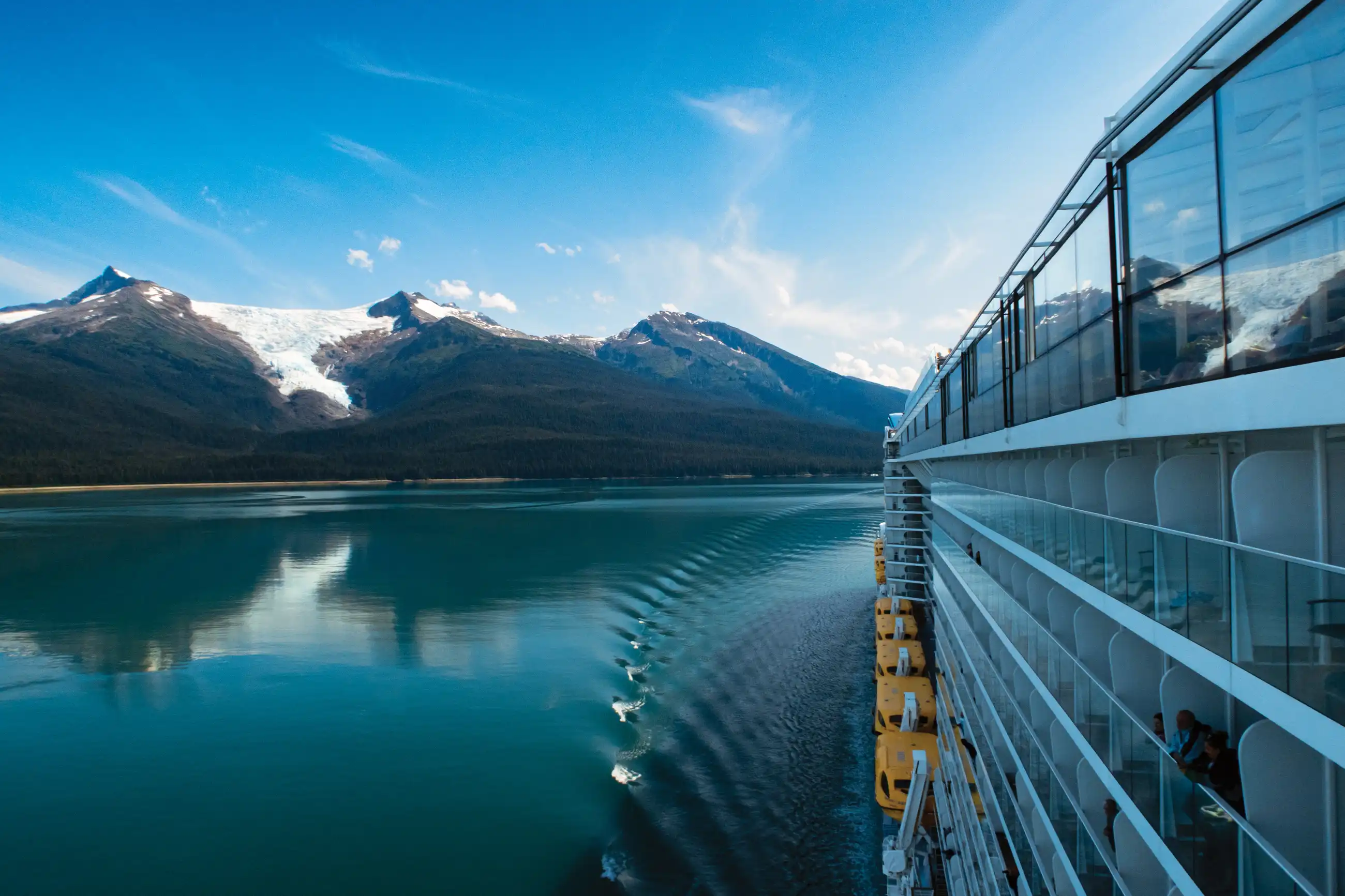 View from the deck railing of Ovation of the Seas across calm turquoise Alaskan water toward snow-capped peaks near Dawes Glacier.
