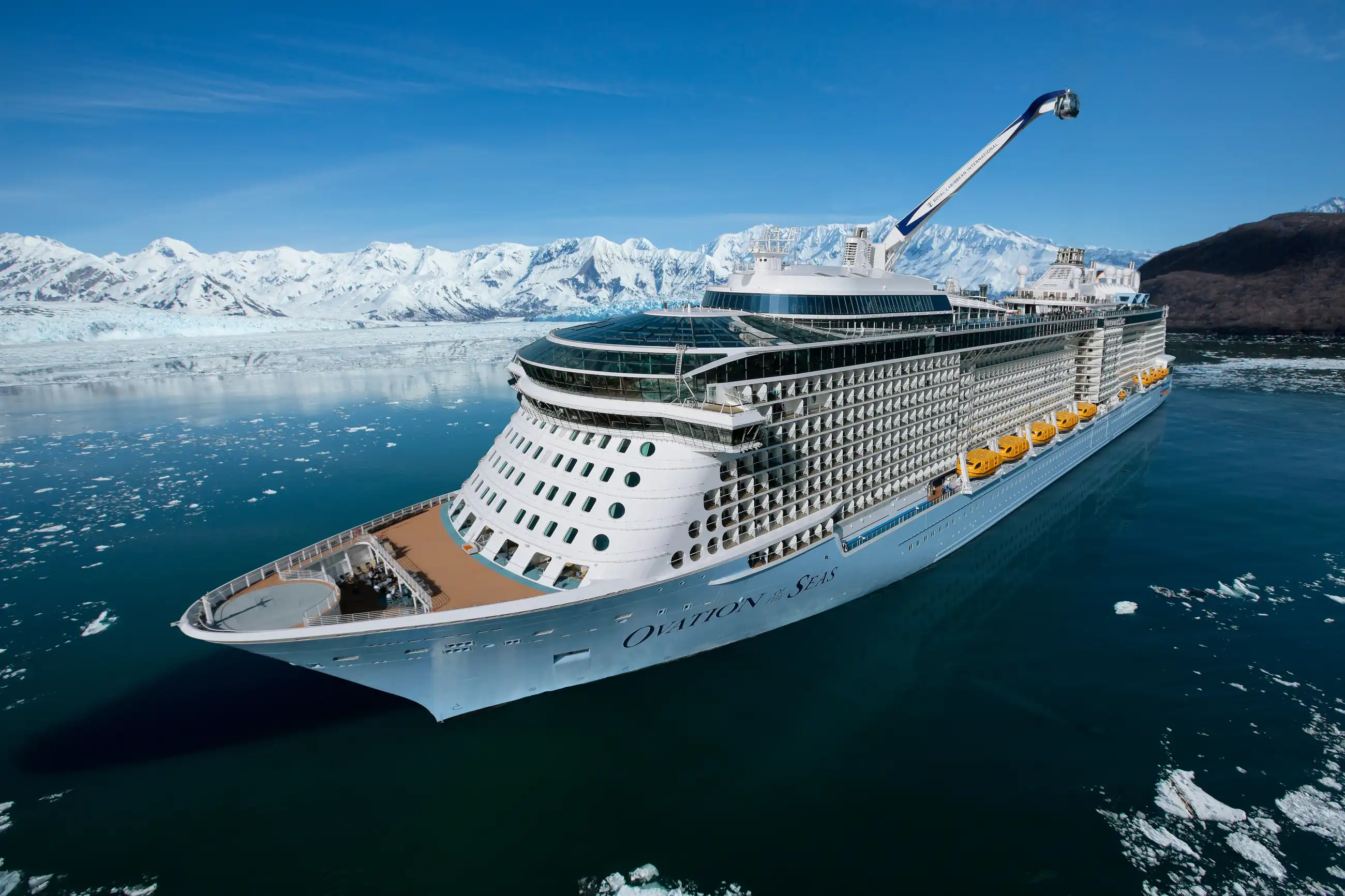 Ovation of the Seas seen in profile in Alaskan waters, surrounded by floating ice with snow-capped mountains behind under clear blue sky.