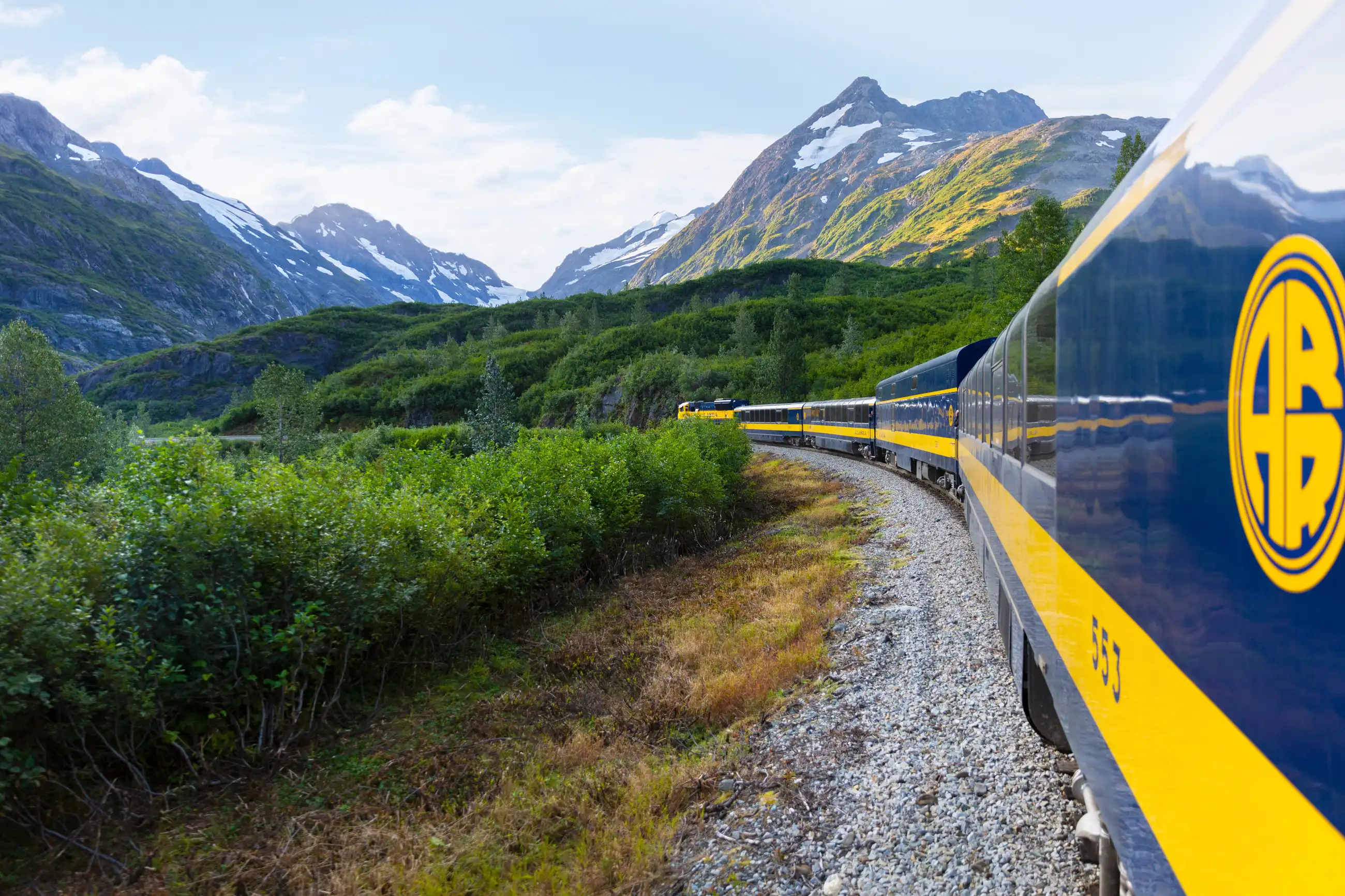 A yellow and blue passenger train runs in profile through a green Alaskan valley, with forested slopes and snow-capped peaks rising behind it under partly cloudy skies.