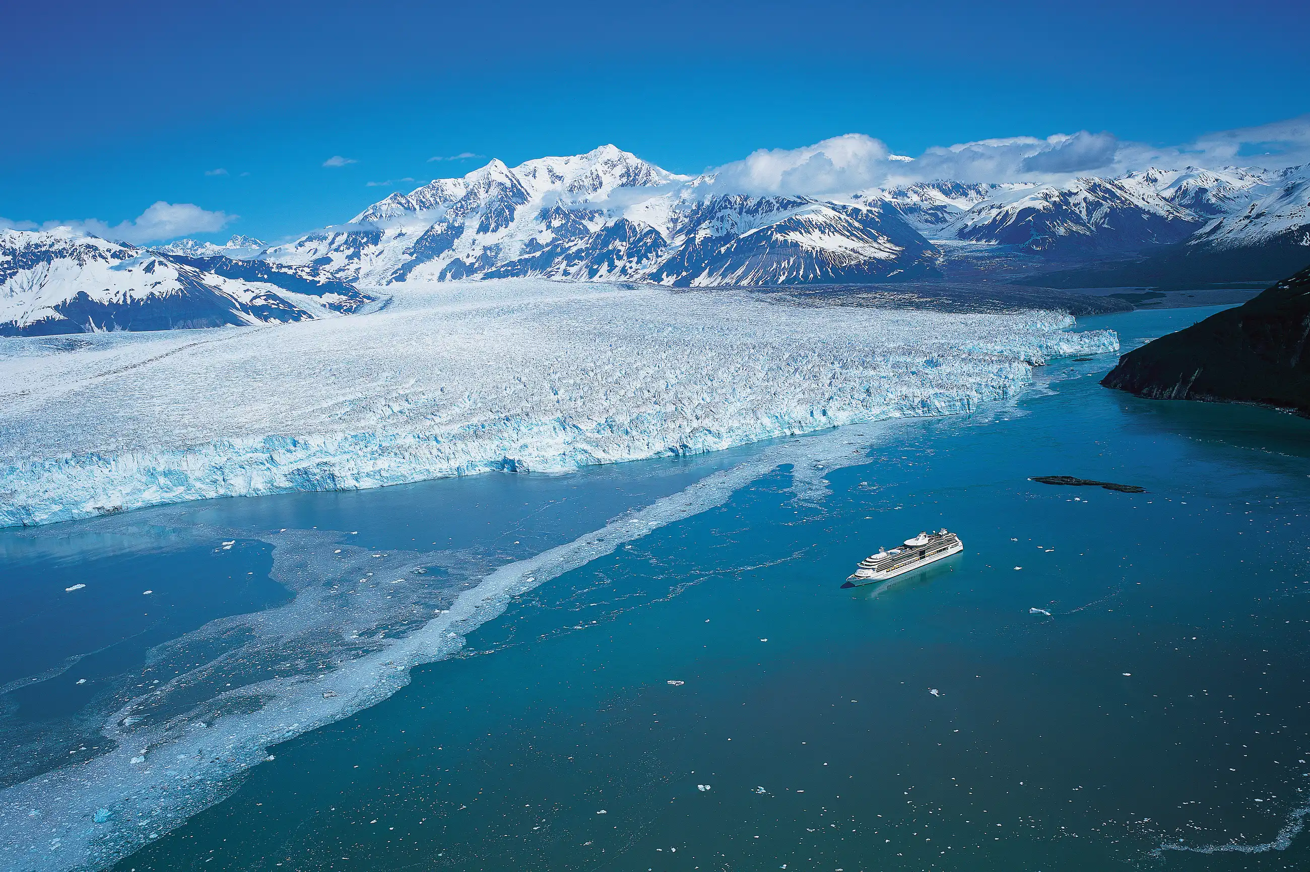 Aerial view of Radiance of the Seas in turquoise glacial water at Hubbard Glacier, Alaska, with the calving glacier face and snow-covered mountains behind.