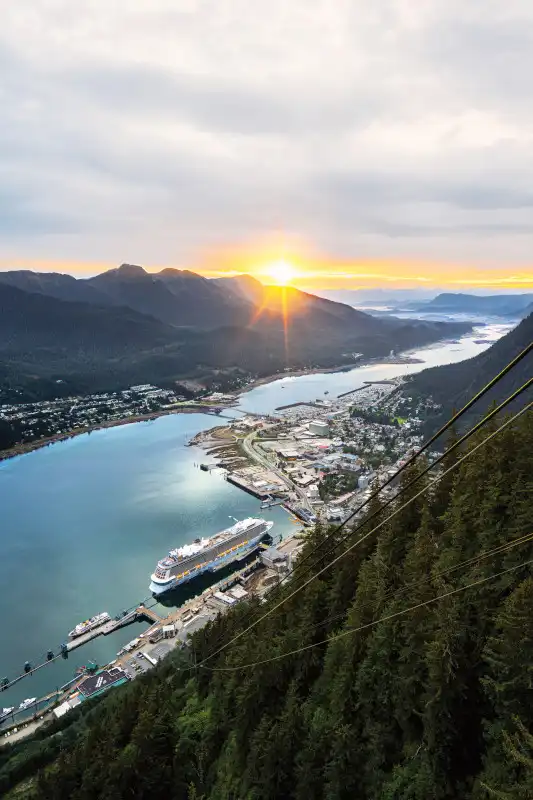 Aerial view taken from a high vantage point overlooking a cruise ship docked in a turquoise-blue harbor, with a mountain valley, forested slopes, and coastal town visible below during golden hour sunset with dramatic sun rays breaking through clouds.