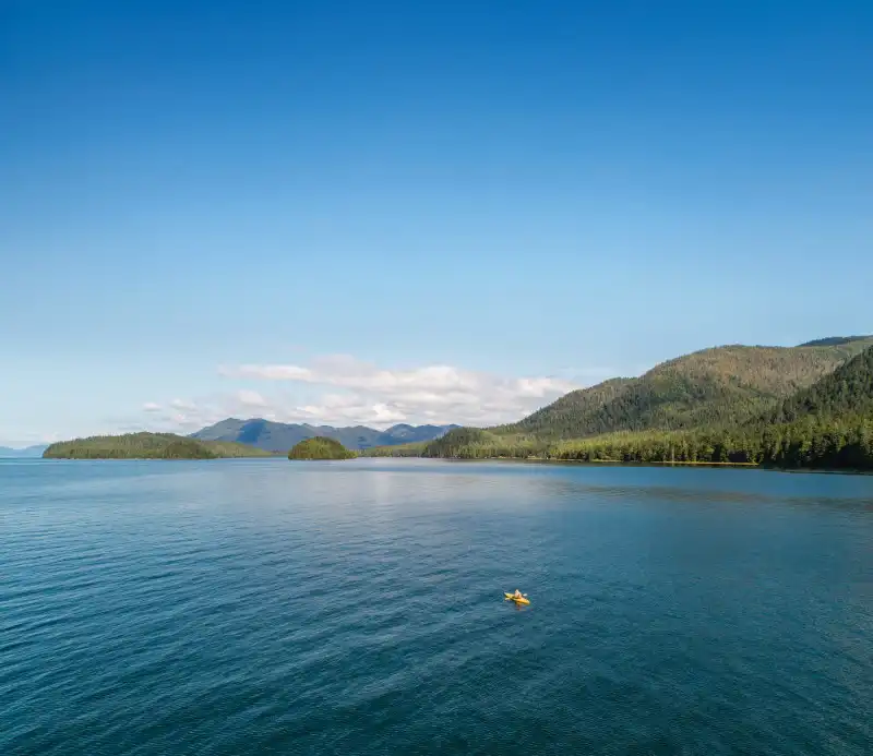 Aerial drone view of a pristine coastal seascape featuring a small yellow kayak on turquoise water, framed by forested shorelines with mountains in the distance under clear blue sky. This scenic Alaska landscape captures the natural beauty of the Inside Passage region.
