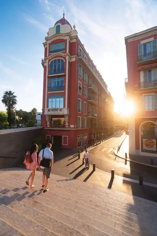 A vibrant red Belle Époque building with a distinctive domed corner tower dominates this Mediterranean port-side street scene. Pedestrians stroll through the sunlit plaza at golden hour, with palm trees and period architecture framing a classic French Riviera setting.