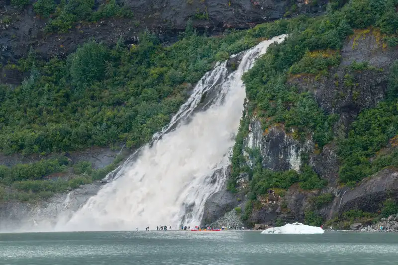 A dramatic cascading waterfall plunges down a steep rocky cliff face surrounded by dense green vegetation into turquoise glacial waters. A small cruise ship tender with passengers is visible at the base, providing scale to this majestic natural feature.