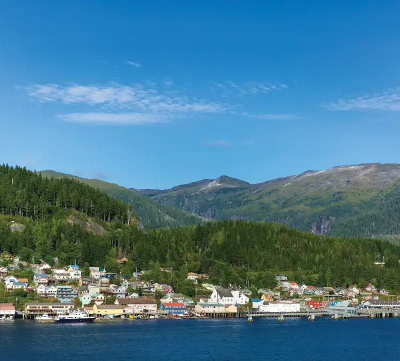 Waterfront view of Ketchikan, Alaska showing a picturesque coastal town nestled against dense coniferous forests with steep mountains rising in the background. The composition captures the settlement's waterfront buildings and docks fronting calm blue waters under clear skies.