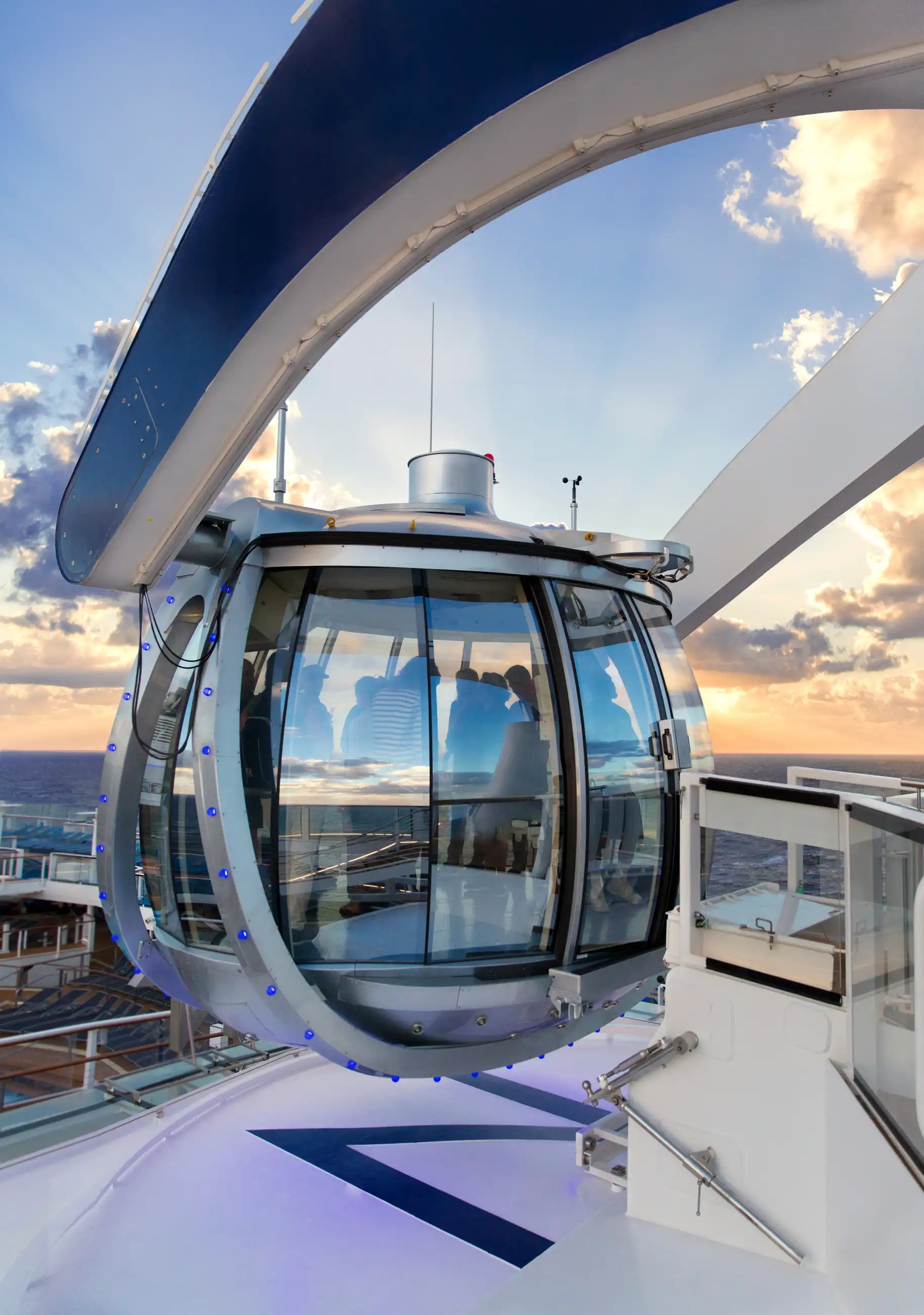 The glass observation capsule on the exterior deck of Anthem of the Seas, silhouetted passengers inside, against a dramatic sunset sky over the ocean.