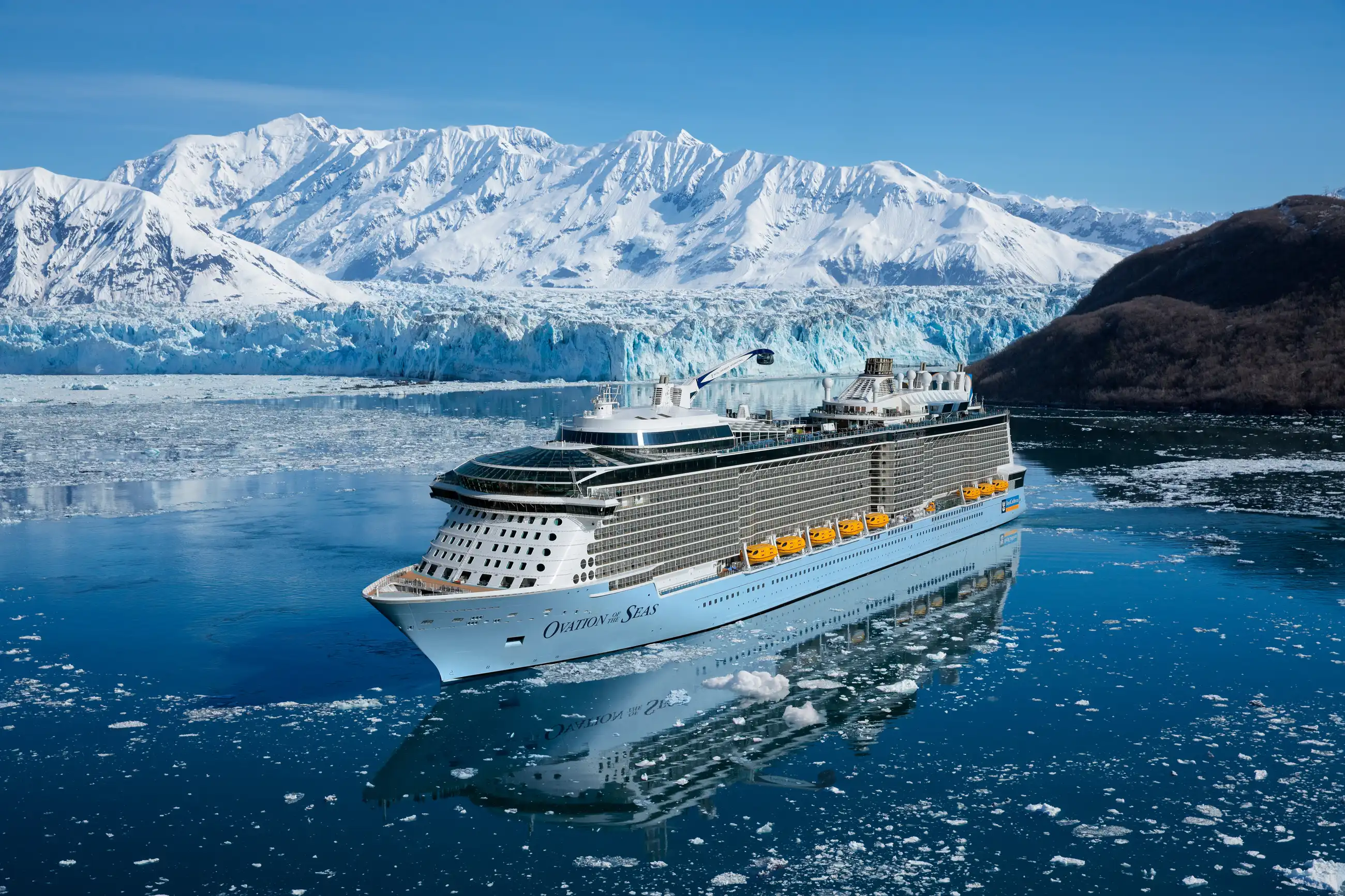 Ovation of the Seas with its yellow lifeboats in icy Alaskan water, with a glacier and snow-covered mountains behind under clear sky.