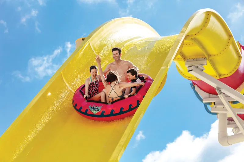 A family of five rides down a bright yellow waterslide on a red inflatable tube in bright daylight. The composition captures the action mid-descent against a clear blue sky with white clouds, with the curved yellow slide structure dominating the frame.