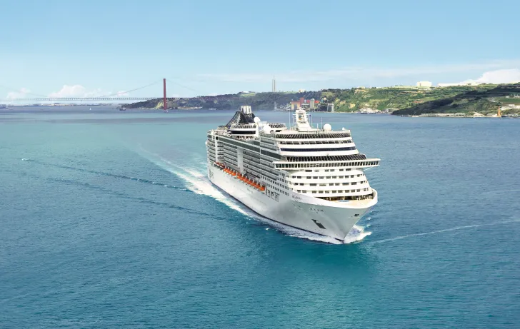 Large modern cruise ship photographed from above in turquoise waters, moving through a Mediterranean or Atlantic location with coastal hills and industrial infrastructure visible in the background. The vessel displays multiple passenger decks with distinctive white livery and orange/red accent bands.