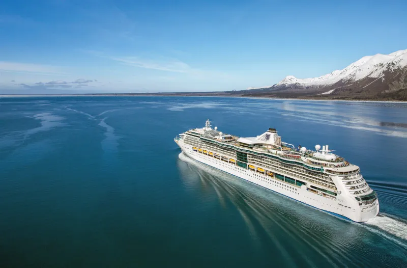 Aerial view of a large cruise ship positioned in calm turquoise waters with snow-covered mountains visible along the coastline. The vessel is photographed from above in clear daylight conditions, showcasing the ship's full profile against pristine natural surroundings.