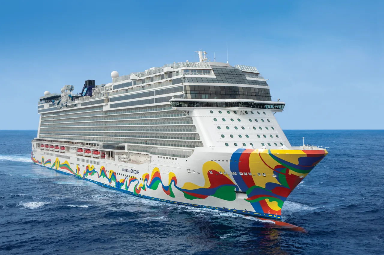 A large modern cruise ship photographed from an aerial vantage point in open ocean. The vessel features distinctive colorful artwork on its white hull with red, yellow, green, and blue wave-like designs, and displays multiple deck levels with rows of portholes against a clear blue sky and calm sea.