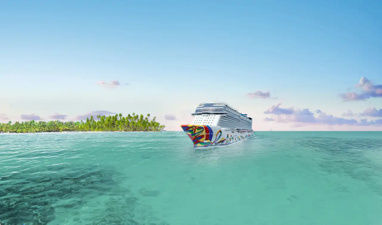 A cruise ship anchored in shallow turquoise water beside a palm-lined Caribbean island at golden hour, with calm seas and scattered cloud.