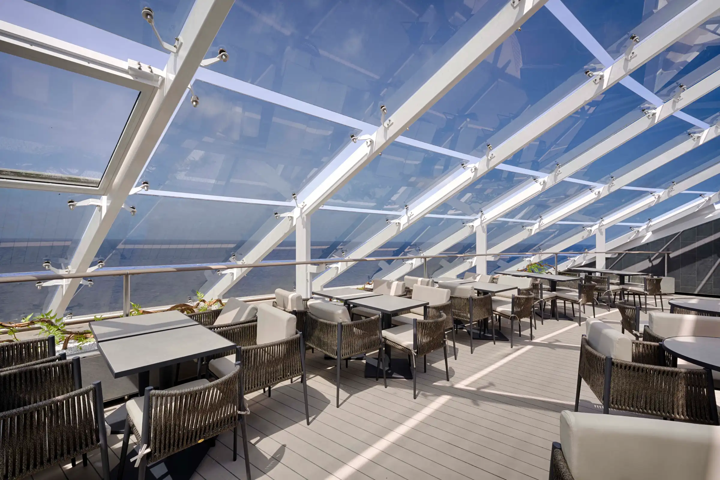 An elegant outdoor dining venue with modern white structural framing supporting a glass ceiling that overlooks open sky and water. The space features contemporary woven chairs, white-topped tables, and potted plants arranged throughout the empty deck area.
