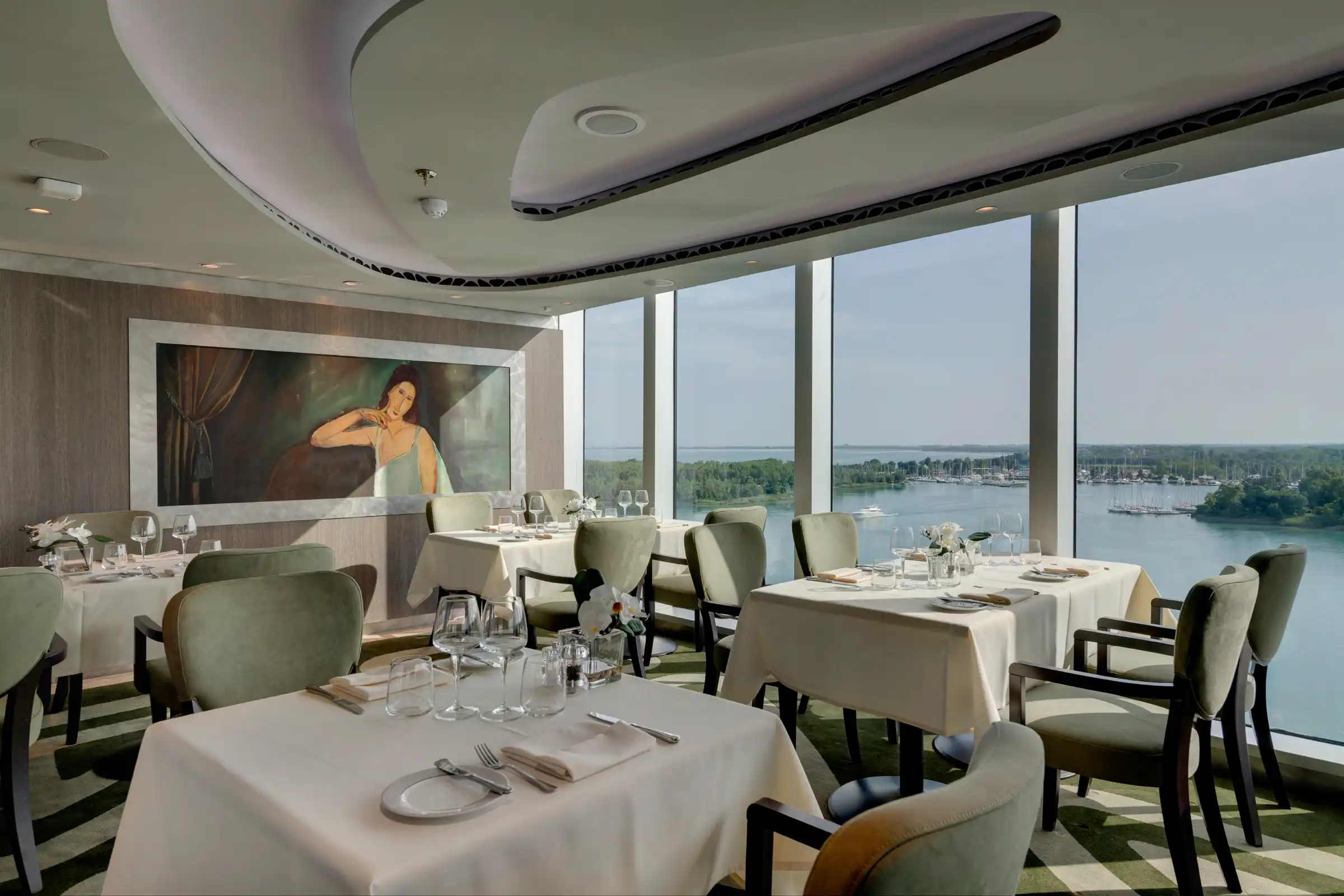 An elegant fine-dining restaurant featuring multiple round tables set with white linens, formal place settings, and upholstered chairs in sage green and black. Floor-to-ceiling windows dominate the composition, offering panoramic views of a waterfront landscape with islands, forests, and a marina visible across calm blue water, while sculptural curved ceiling elements and contemporary artwork complete the sophisticated interior design.
