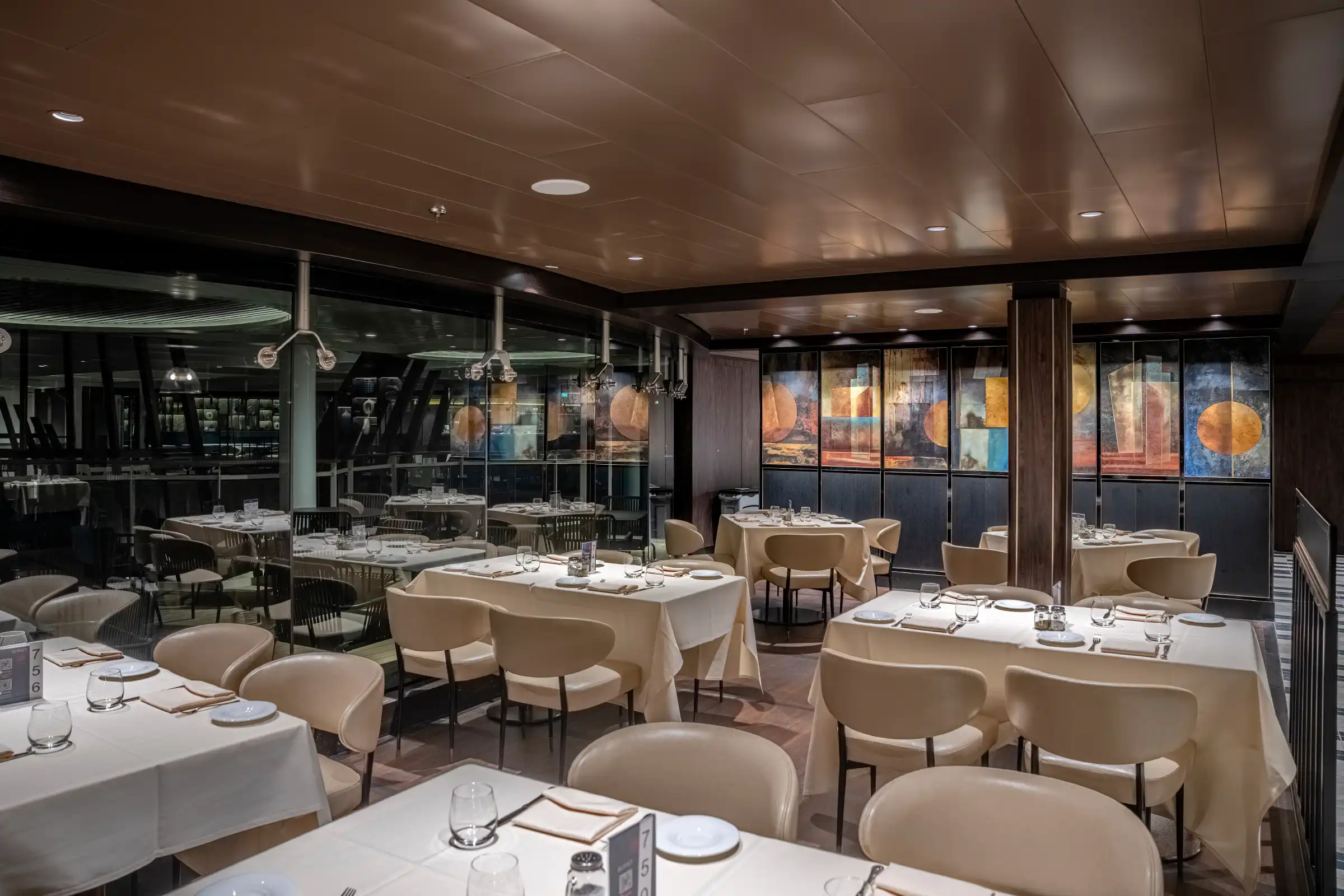 An elegant dining room set for formal dinner service with multiple round tables dressed in cream linens, beige upholstered chairs, and place settings. Floor-to-ceiling windows with illuminated artwork panels frame the venue, while bronze ceiling panels and recessed lighting create an upscale atmosphere.